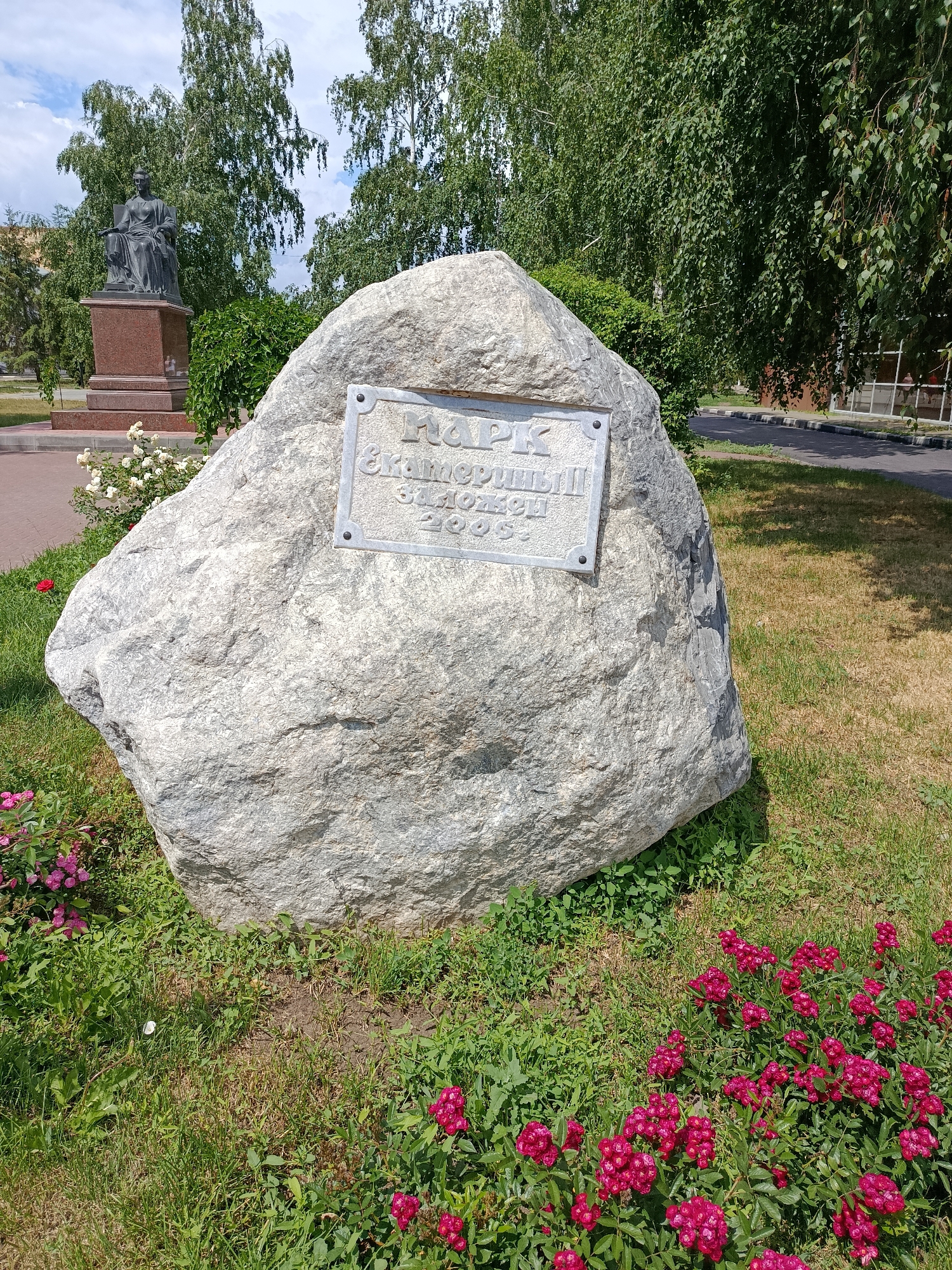 A large, light-gray boulder with an inscribed plaque is the central focus of the image. The plaque, made of a lighter material and secured with four rivets, bears Cyrillic text. The inscription appears to read "ПАРК" at the top, followed by "Екатериненный" and "1909", suggesting the park's name and founding year. The boulder is situated on a grassy area, with patches of dry grass and some green shoots. Pink flowers are blooming in clusters around the base of the boulder in the foreground. In the background, to the left, is a dark bronze statue of a seated figure on a red granite pedestal, partially obscured by greenery. Tall, leafy trees with light bark, likely birch trees, dominate the background, their branches creating a dappled shade. A paved walkway with a black and white striped border is visible to the right, suggesting a public park or garden setting in the city of Marx, Russia. The sky is partly cloudy with patches of blue.