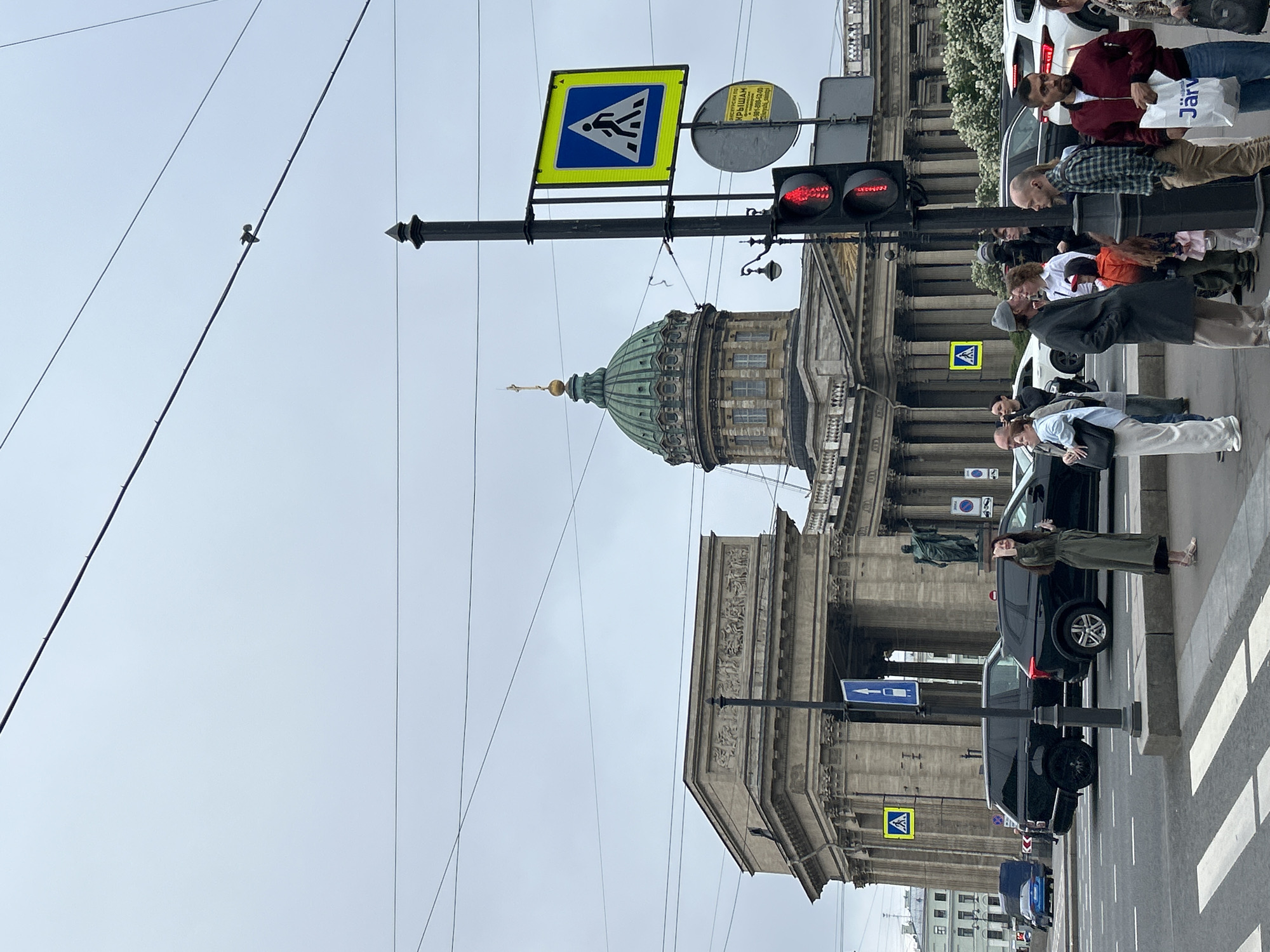 The image depicts a street scene in Saint Petersburg, Russia, focusing on the Kazan Cathedral. The cathedral is a prominent feature, with its large dome and architectural details visible in the background. In the foreground, there's a pedestrian crosswalk sign with a pedestrian symbol in blue on a yellow background, a traffic light showing red, and a group of people waiting to cross the street. People are standing on the sidewalk, dressed in various casual clothing. Cars are parked on the street, and some are moving. The sky is overcast, and power lines are visible stretching across the top left of the frame. The overall scene is a typical urban environment with pedestrians, traffic, and iconic architecture.