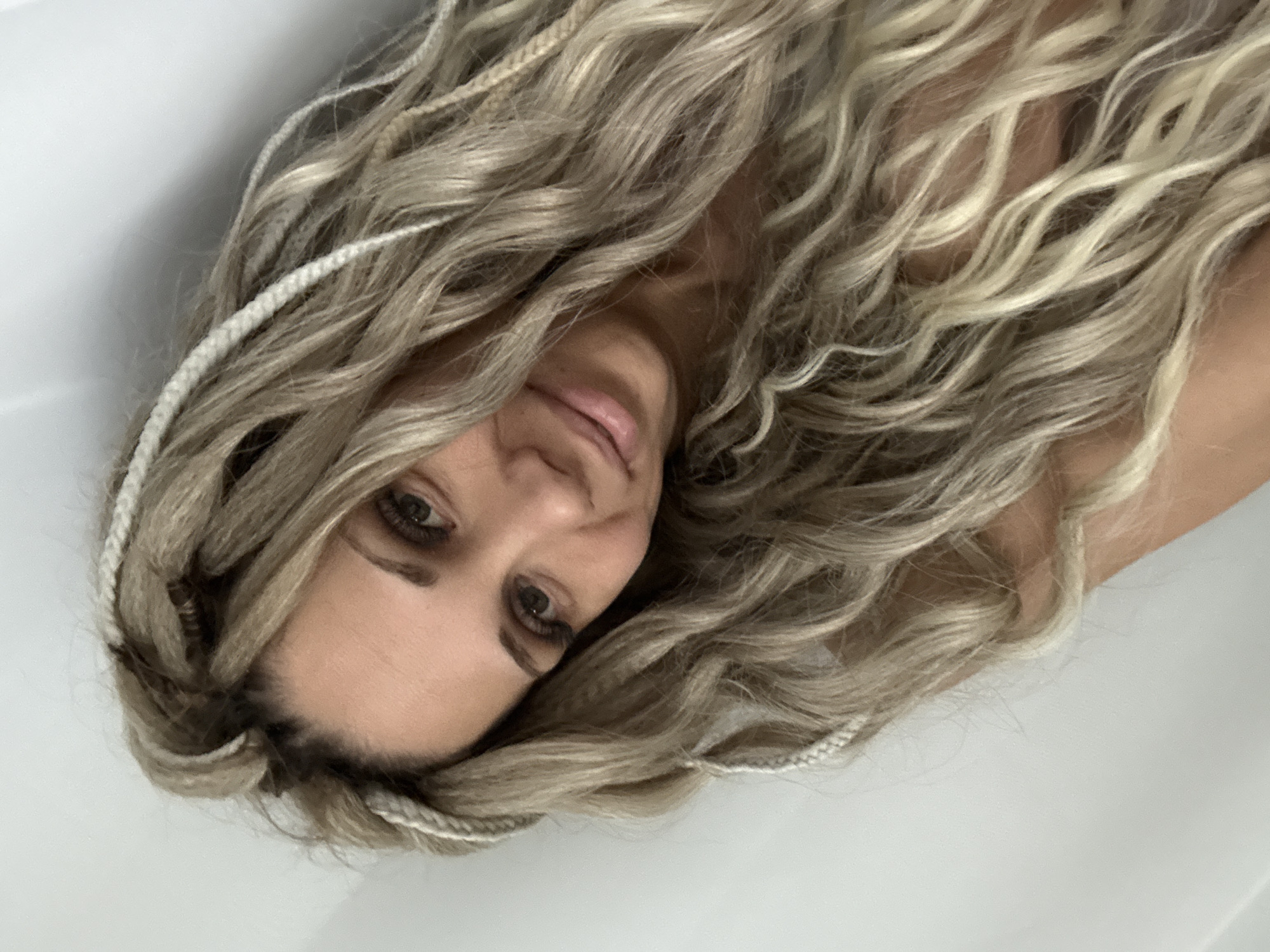 A woman with blonde wavy hair and light-colored braided accents is lying in a white bathtub. Her face is turned towards the camera. The environment suggests a private residence in an unspecified location; there is no evidence indicating Rusanovo, Russia.