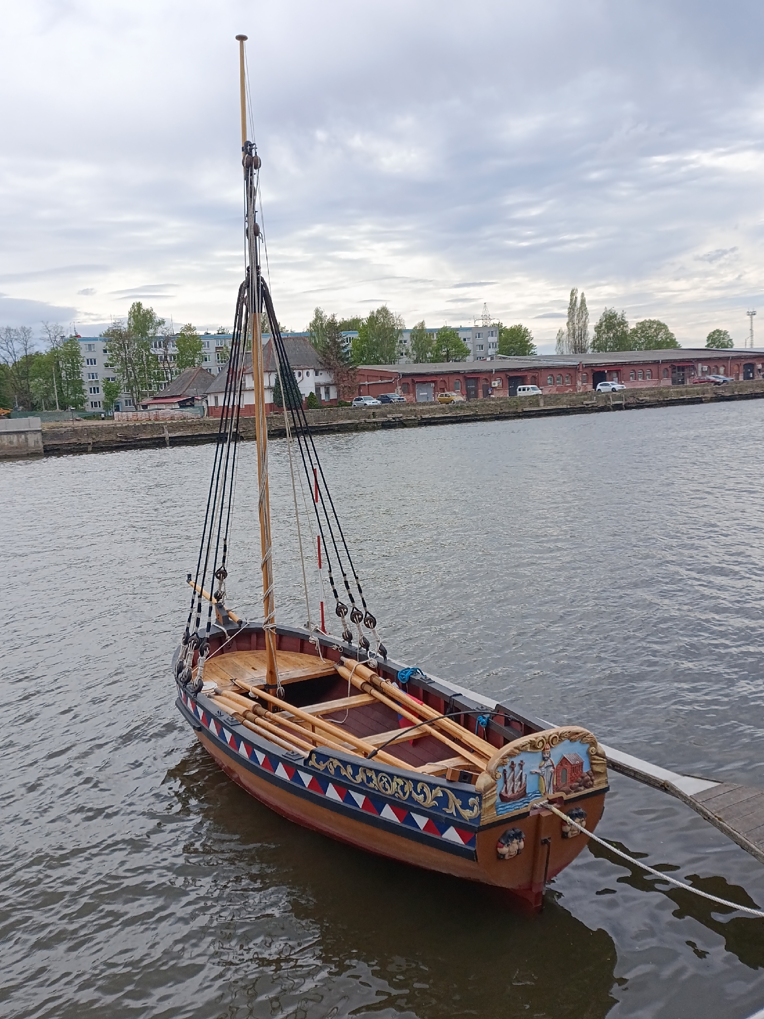 A high-angle medium shot depicts a small, vintage sailboat tethered at a dock in Kaliningrad, Russia. The boat, with its wooden hull, is painted with a combination of brown, red, blue, and white colors and has decorative, figurative carvings near the stern. The mast is a tall wooden pole adorned with ropes and pulleys. Several oars are placed inside the boat. The boat floats in water, which shows ripples. In the background, across the water, stands a building with multiple windows and red-brick sections. Several cars are parked along the street in front of the building. A cloudy sky fills the upper part of the frame. The location is identifiable as Kaliningrad due to the style of the boat, the architecture of the buildings, and common tourist attractions.