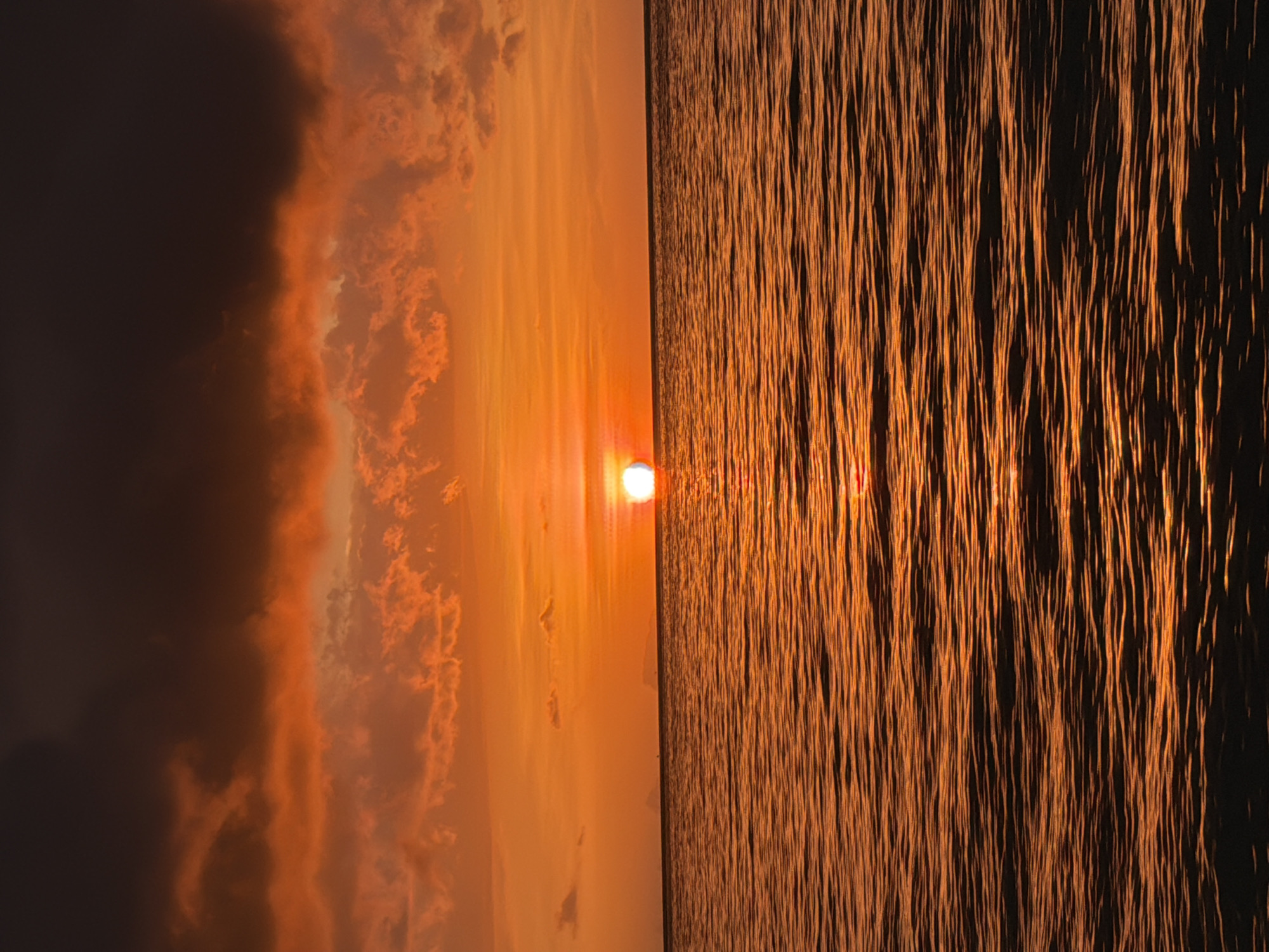The image captures a sunset over the ocean in Baan Tai, Thailand. The sun, a bright white orb, is low on the horizon, casting a fiery orange glow across the sky and the rippling water. Dark, dramatic clouds dominate the upper portion of the frame, contrasting with the vibrant hues of the sunset. The ocean's surface is textured with numerous small waves, reflecting the warm light and creating a shimmering effect. The scene is devoid of any human presence or man-made structures, focusing solely on the natural spectacle of the sunset.