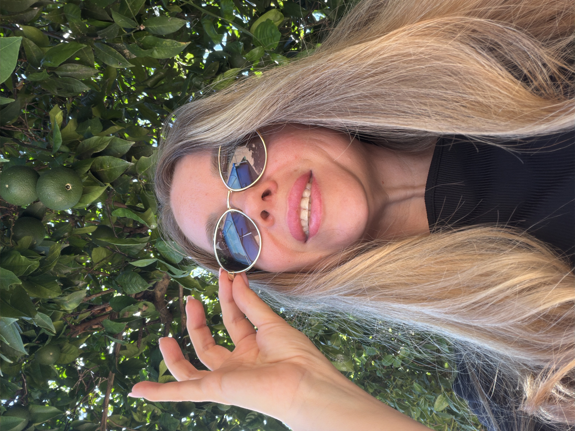 A close-up, low-angle outdoor selfie shows a woman with long, blonde hair and sunglasses in front of a backdrop of green leaves and branches, with some unripe green fruit visible. She is wearing a black, ribbed top and is holding her sunglasses up with her left hand, her fingers partially obscuring the lenses. Her mouth is open in a slight smile, revealing her teeth. The sunglasses reflect a blue object, possibly a building or sky, and possibly a portion of a blue swimming pool. The image appears to have been taken in Rome, Italy, based on contextual information.