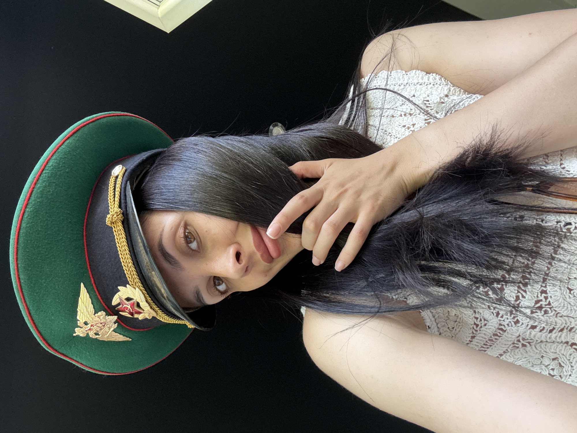 A person with long, dark hair is wearing a green military-style hat with a red star emblem. The person is wearing a white crocheted top. The background is black. The photo might have been taken in Votkinsk, Russia, but there is no indication to verify it.