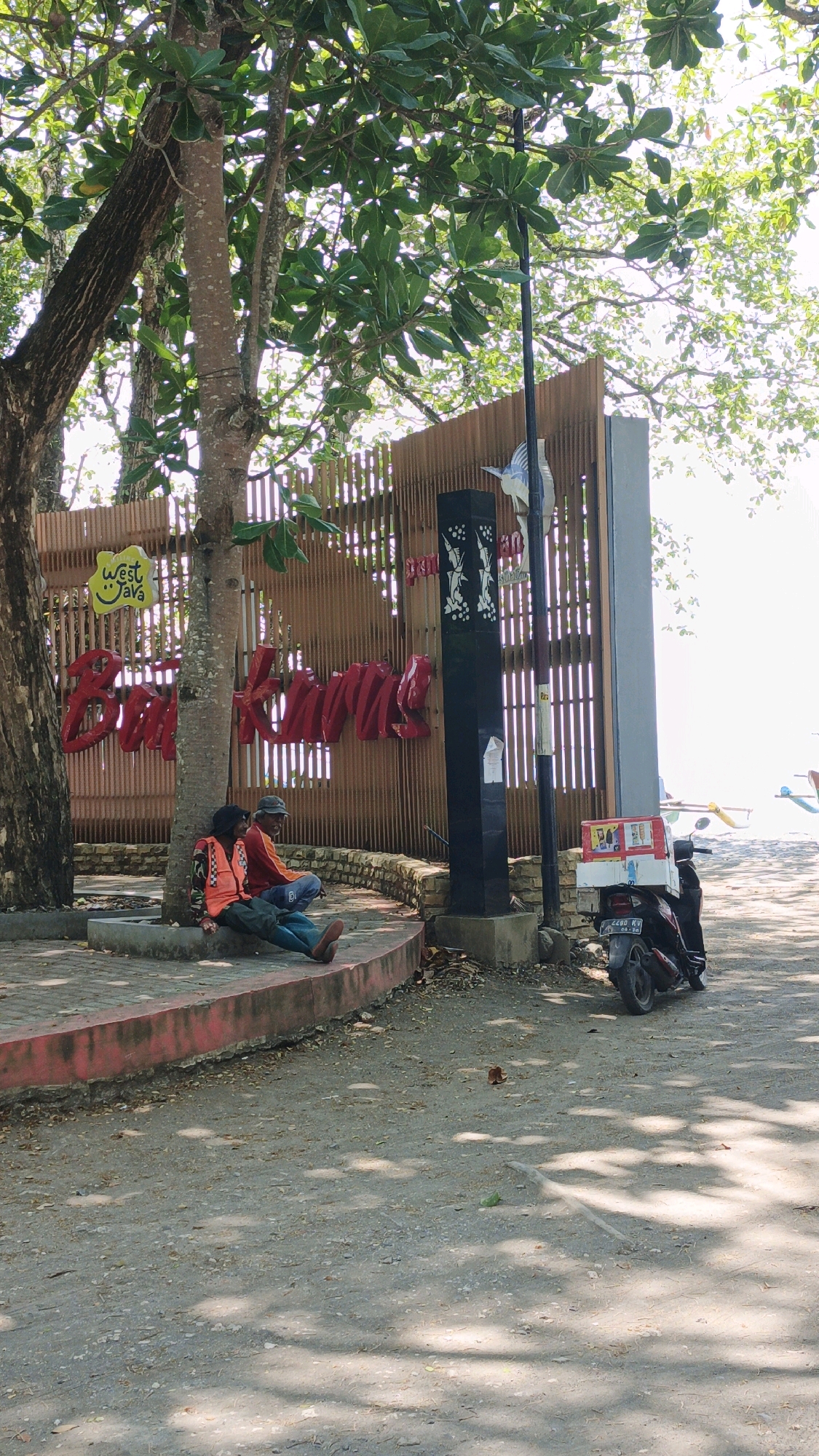 Two men are sitting on a curved concrete ledge next to a tree in Desa Batukaras, Indonesia. The man on the left is wearing a green shirt, an orange life vest, and dark pants. He is leaning against the tree with his legs extended. The man on the right is wearing an orange shirt and a grey baseball cap. He is sitting with his legs crossed and looking towards the left. Behind them is a wooden fence with a large red sign that reads "Batik Keras" and a smaller yellow sign with the words "West Java". A black, rectangular pillar with white decorative elements stands to the right of the sign. A motorcycle with a white box on the back is parked on the dirt road to the right of the pillar. The environment appears to be outdoors, with trees providing shade over the ground. In the background, there is a body of water, possibly the sea, and a boat.