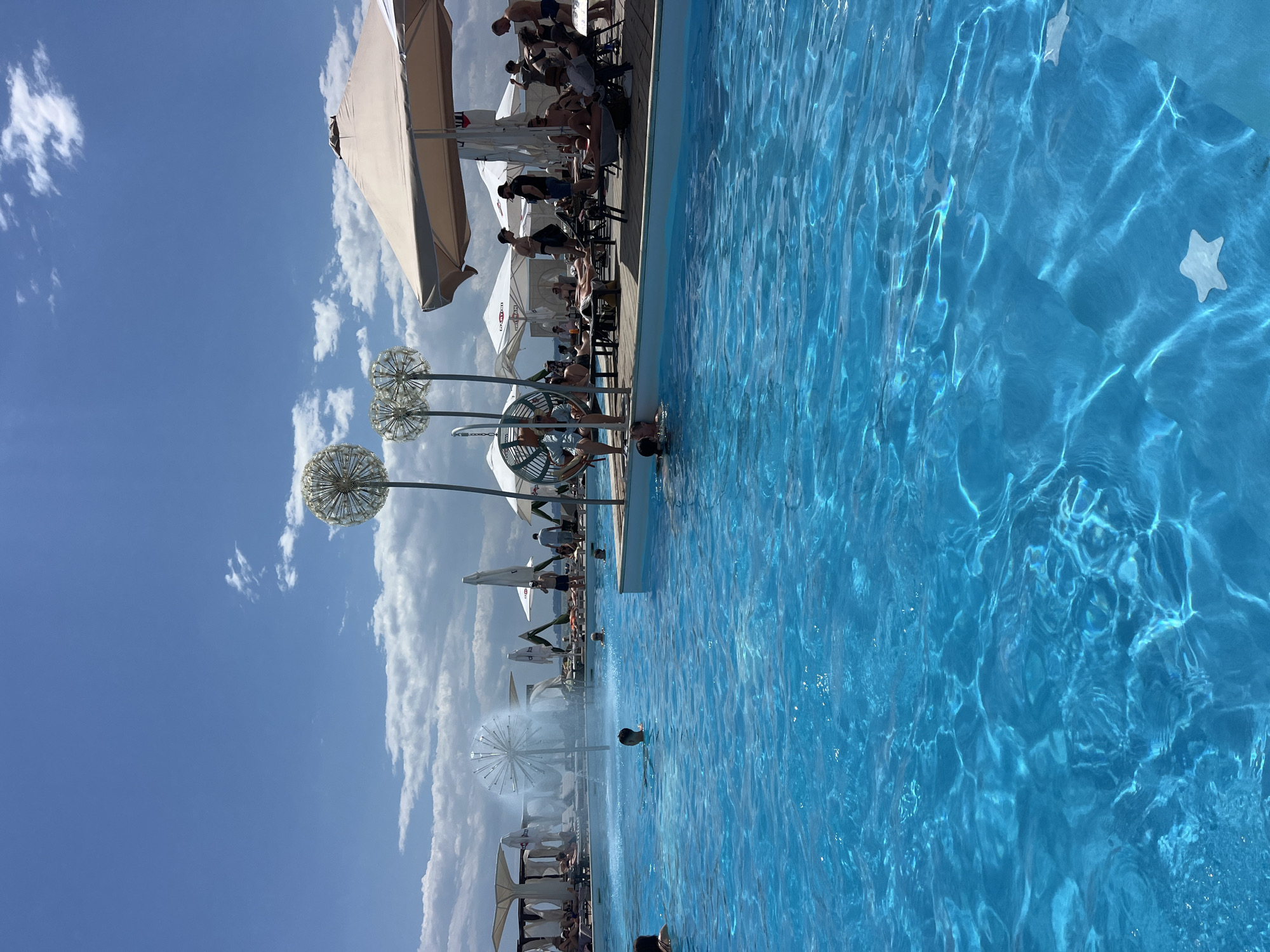 This is a daytime photograph taken at a pool club on the coast of Fontanka, Ukraine. The scene is dominated by a vibrant, bright blue swimming pool filled with clear water, reflecting sunlight and creating shimmering patterns on the surface. In the foreground of the pool, two white star-shaped floaties are visible. Several people are enjoying the water, with some swimming and others partially submerged.
The background of the image reveals a bustling pool club setting. Along the edge of the pool, there is a wooden deck where lounge chairs and umbrellas are set up. Numerous individuals are seated or lying on the lounge chairs, relaxing under the shade of the umbrellas. Some are dressed in swimwear, while others are in casual clothing. A fountain is active in the pool, spraying water upwards. Decorative, spherical, dandelion-like sculptures stand near the pool, adding an artistic element to the environment. In the distance, more umbrellas and structures suggest a larger resort or entertainment complex. The sky above is a clear, bright blue with scattered white clouds. The overall atmosphere appears to be one of leisure and enjoyment on a sunny day.