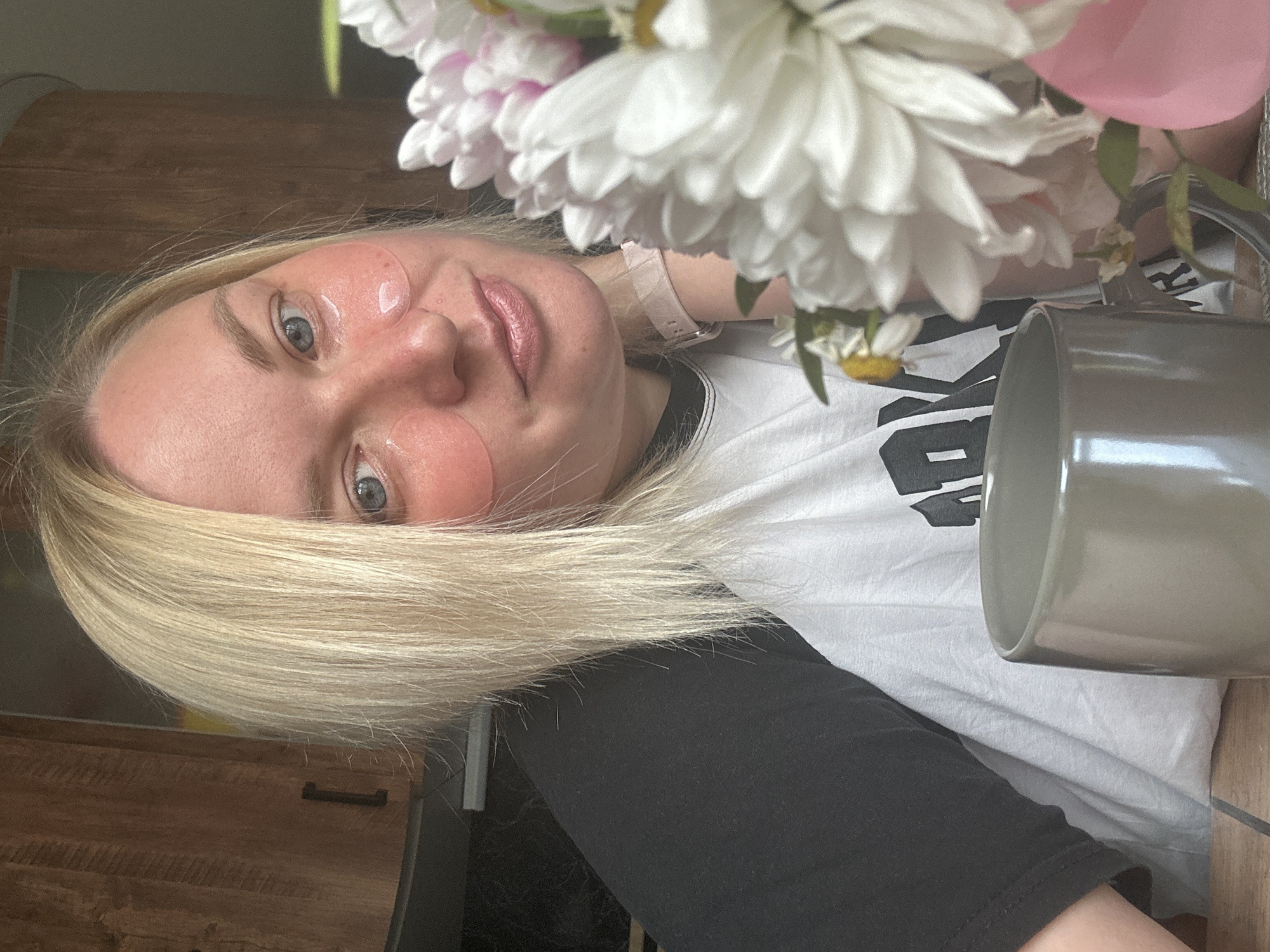 The image shows a close-up selfie of a woman with blonde hair, blue eyes, and pink under-eye patches. She is wearing a black and white t-shirt. In the foreground, there is a grey mug and a bouquet of white and pink flowers. The background appears to be a kitchen with wooden cabinets. The woman is leaning her head on her hand, and her facial expression is neutral. There is no visible indication of the location being Chaikovsky, Russia.