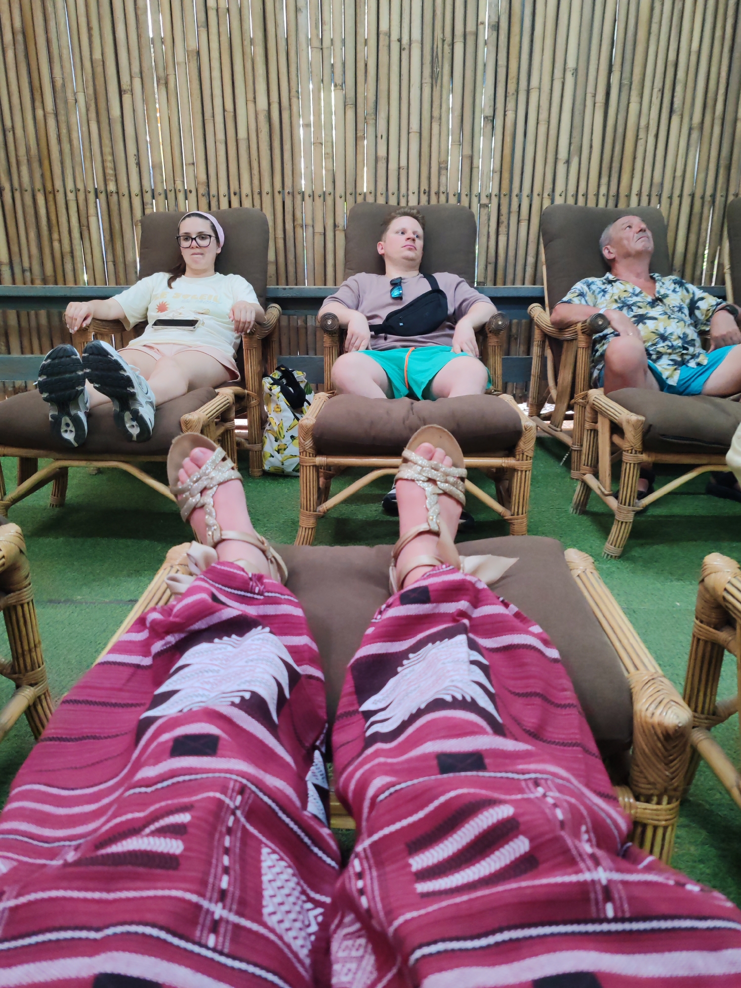 This media file captures a first-person perspective of people relaxing in a setting suggestive of Ban Khlong Khian, Thailand. The foreground shows the lower legs and feet of the person taking the photo, adorned in gold-colored sandals and vibrant red pants or a sarong with white and black geometric patterns.
Three other individuals are visible, seated in dark brown cushioned rattan chairs arranged in a line against a textured bamboo wall. From left to right:
1. A woman with dark hair pulled back, wearing a light pink headband, glasses, a light yellow t-shirt, and light-colored shorts. Her white and black sneakers are resting on the chair's footrest. She looks directly towards the camera with a neutral expression, a mobile phone resting on her lap.
2. A younger man with short blonde hair, wearing a light purple t-shirt and teal shorts. A black fanny pack is slung across his chest, and sunglasses rest on his shirt collar. He looks upwards and to his left.
3. An older man with short grey hair, wearing a floral short-sleeved button-up shirt with blue and yellow patterns, and light blue shorts. He looks upwards and to his right.
The chairs are made of light brown rattan, and the floor beneath them appears to be green artificial turf. A yellow and white banana-patterned bag is placed between the first and second chairs. The bamboo wall in the background provides a natural, tropical aesthetic, with bright light filtering through its gaps, suggesting daytime. The overall atmosphere is one of relaxation and leisure. No discernable text is visible on any objects or clothing.