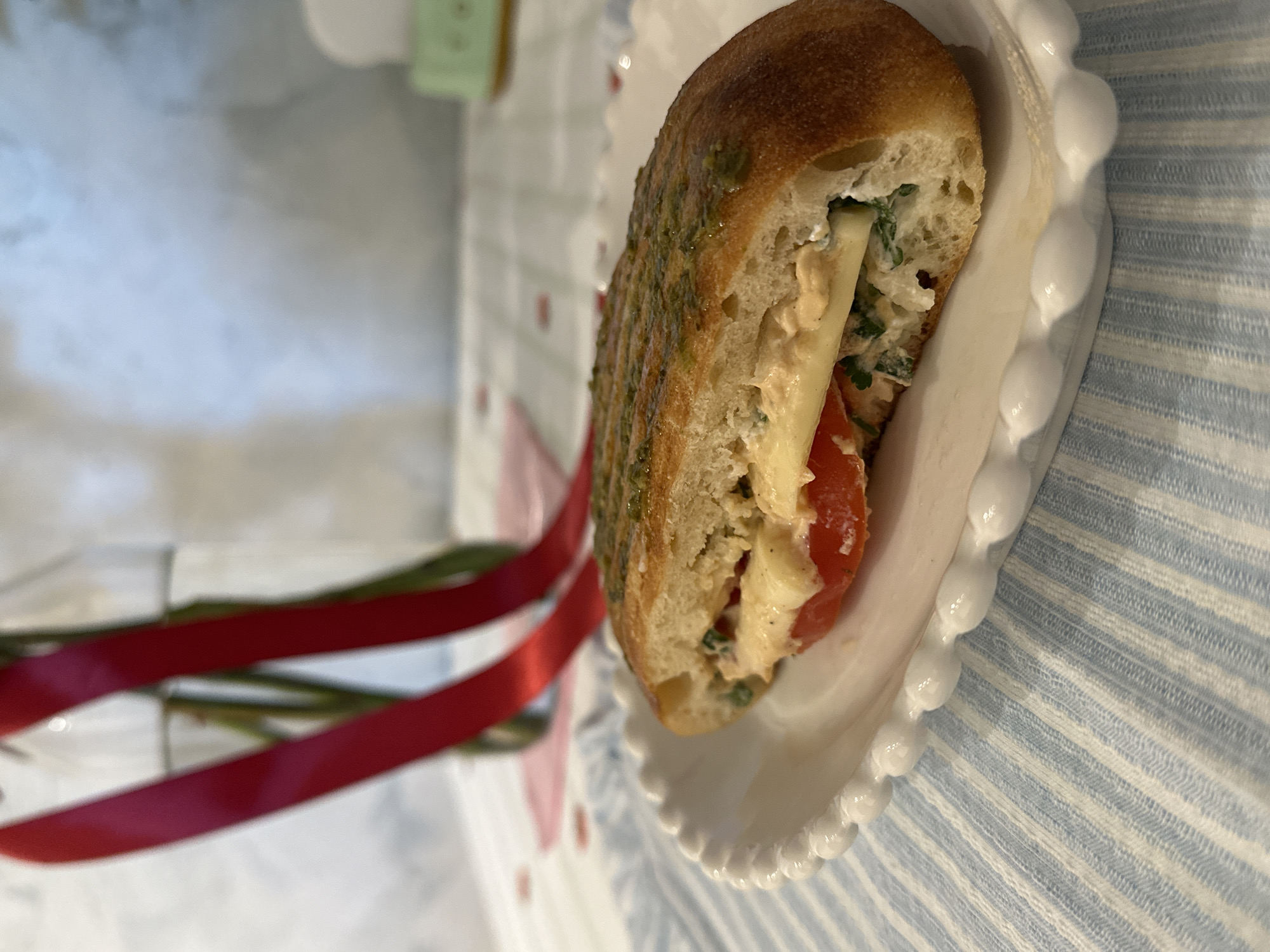 A close-up shot depicts a sandwich on a white, ornate plate. The sandwich contains sliced tomatoes, cheese, and a mixture of greens and creamy filling between two halves of bread, with a green topping. The plate rests on a blue and white striped tablecloth. In the blurred background, there is a red ribbon or other decoration and a neutral-colored surface. There is no evidence to suggest that this picture was taken in Šušanj, Montenegro.