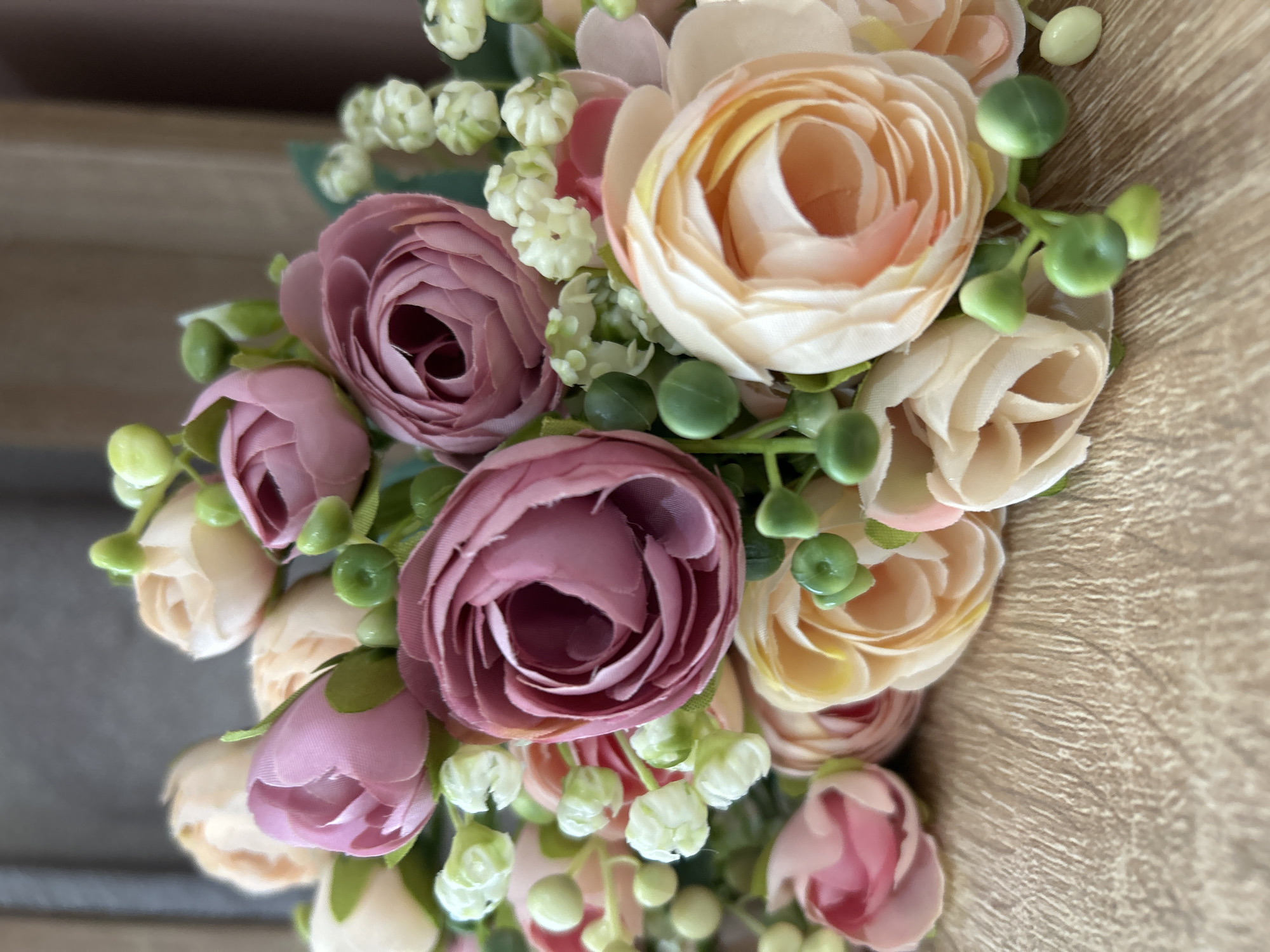The image shows a close-up of a bouquet of artificial flowers. The flowers are various shades of pink and peach, with green buds and small white flowers interspersed. The background is a blurred wooden surface. There is no indication of the location in Chaikovsky, Russia, or any people, actions, or interactions.