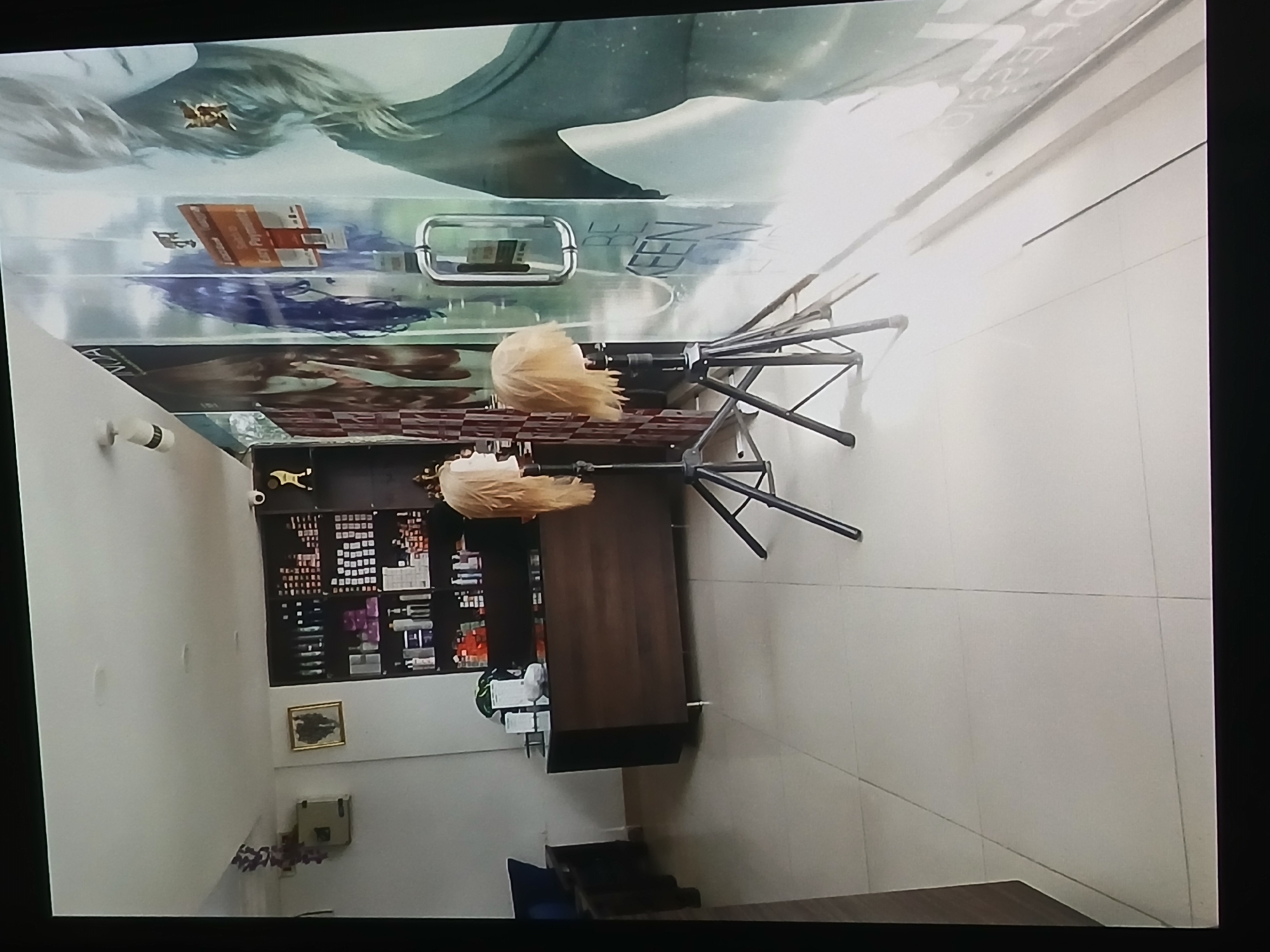 The image depicts the interior of a hair salon in Bengaluru, India. A wall-mounted display features images of models with stylized hair, including one with blonde hair. A reception desk is present with a chair placed behind it. A shelf unit displays numerous haircare products. Surveillance cameras are installed. The floor is tiled. Seating is placed in one of the room's corners. The overall perspective is from above, oriented on its side.