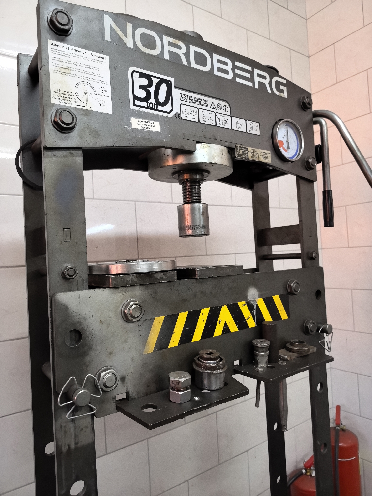 The image shows a NORDGERG 30-ton hydraulic press. The press is made of dark gray metal and is located indoors, with a white tiled wall in the background. A pressure gauge is visible on the right side of the press. There are several labels and stickers on the press, including a warning label in multiple languages, a sticker with the number "30" and "ton", and a serial number sticker. There are also some tools and parts on a shelf attached to the press. A red fire extinguisher is visible in the lower right corner of the image.