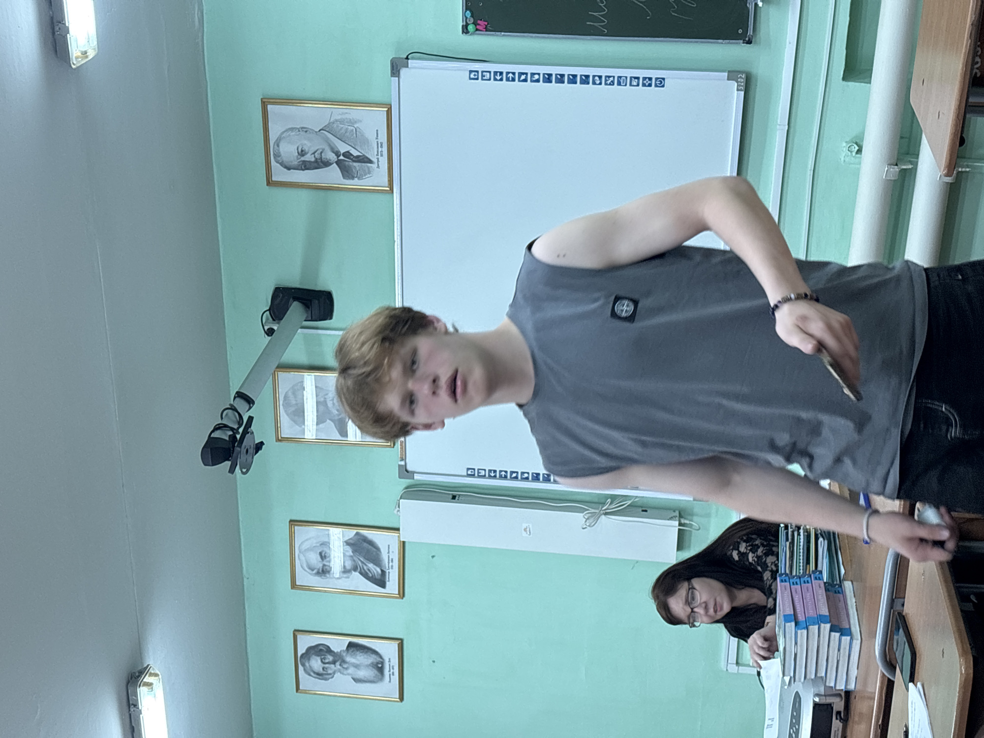 The image captures an indoor scene, likely a classroom or lecture hall, characterized by light green walls and bright fluorescent lighting. The photo is oriented ninety degrees counter-clockwise.
The primary subject is a young male, possibly a teenager, with light brown hair, standing and looking towards the camera. He wears a grey t-shirt with a circular dark patch (possibly a logo) on his left sleeve, and a beaded bracelet on his right wrist. His mouth is slightly open, suggesting he might be speaking or caught mid-sentence.
In the background, to the right of the young man, a woman with dark hair and glasses is seated at a wooden desk, looking down. A tall stack of books with purple and white spines, displaying the Cyrillic word "ЛИТЕРАТУРА" (Literature), rests on the desk beside her, along with what appears to be a mobile phone.
The wall features several framed, black-and-white portraits of historical figures. One clearly identifiable portrait is of "Дмитрий Иванович Менделеев" (Dmitri Ivanovich Mendeleev), with dates "1834-1907". Another portrait, partially obscured, might be of Alexander Pushkin. Some portraits have a horizontal white band across the eyes, possibly a stylistic or modified element. A whiteboard with a row of function icons (e.g., undo, redo, save, zoom) is mounted on the wall, with a traditional blackboard visible above it, showing some illegible white chalk writing. A projector arm or camera mount extends from the wall towards the center of the room.
The setting, with its portraits, white/blackboards, and textbooks, strongly suggests an educational environment in a Russian-speaking country. The time of day is not discernible, but the reliance on artificial lighting indicates it could be any time of day.