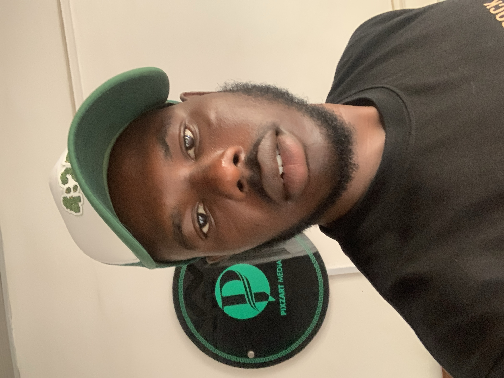A man is featured in a close-up, chest-up shot, appearing to take a selfie or be photographed. He has dark skin, a beard, and is looking directly at the camera with a neutral to slightly smiling expression. He is wearing a black crew-neck t-shirt, with a partially visible "OCK" text on the right shoulder. On his head, he wears a green and white baseball cap with a green brim and a white front panel that has a small green, stylized logo or patch.
In the background, a light-colored wall is visible. To the man's lower left (when the image is viewed upright), a circular black sign with a prominent green logo and text is mounted on the wall. The logo consists of stylized intertwined shapes, and the text clearly reads "PIXZART MEDIA". The sign also features a green border with a repeating pattern. The setting appears to be indoors, possibly an office or commercial space, in Jos, Nigeria.