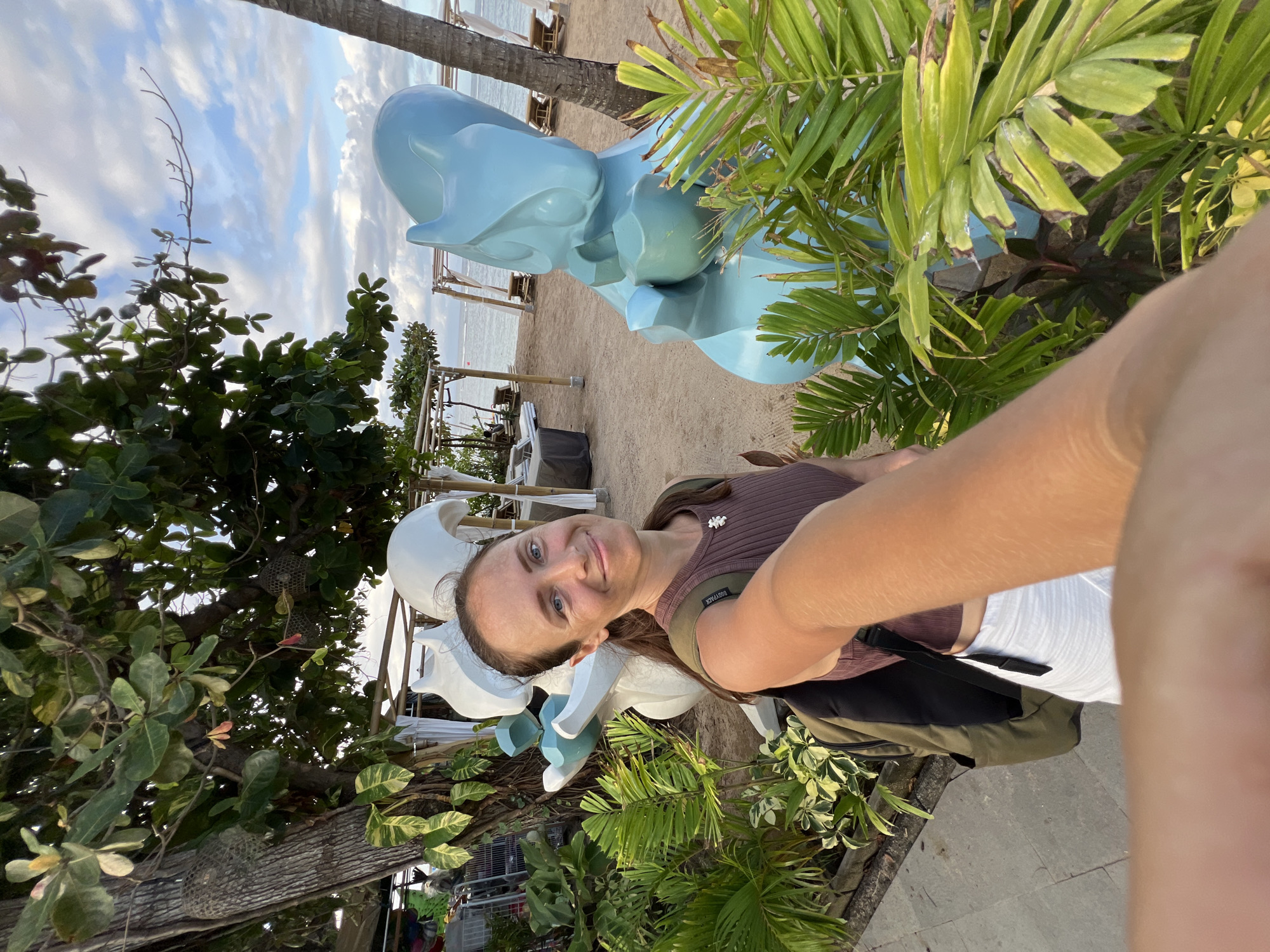 A woman with brown hair and blue eyes is taking a selfie at the beach in Denpasar, Indonesia. She is wearing a brown shirt, white shorts, and a backpack. Behind her, there is a light blue dolphin sculpture and various tropical plants. In the background, a sandy beach leads to the ocean under a cloudy sky. A few wooden structures are visible on the beach.