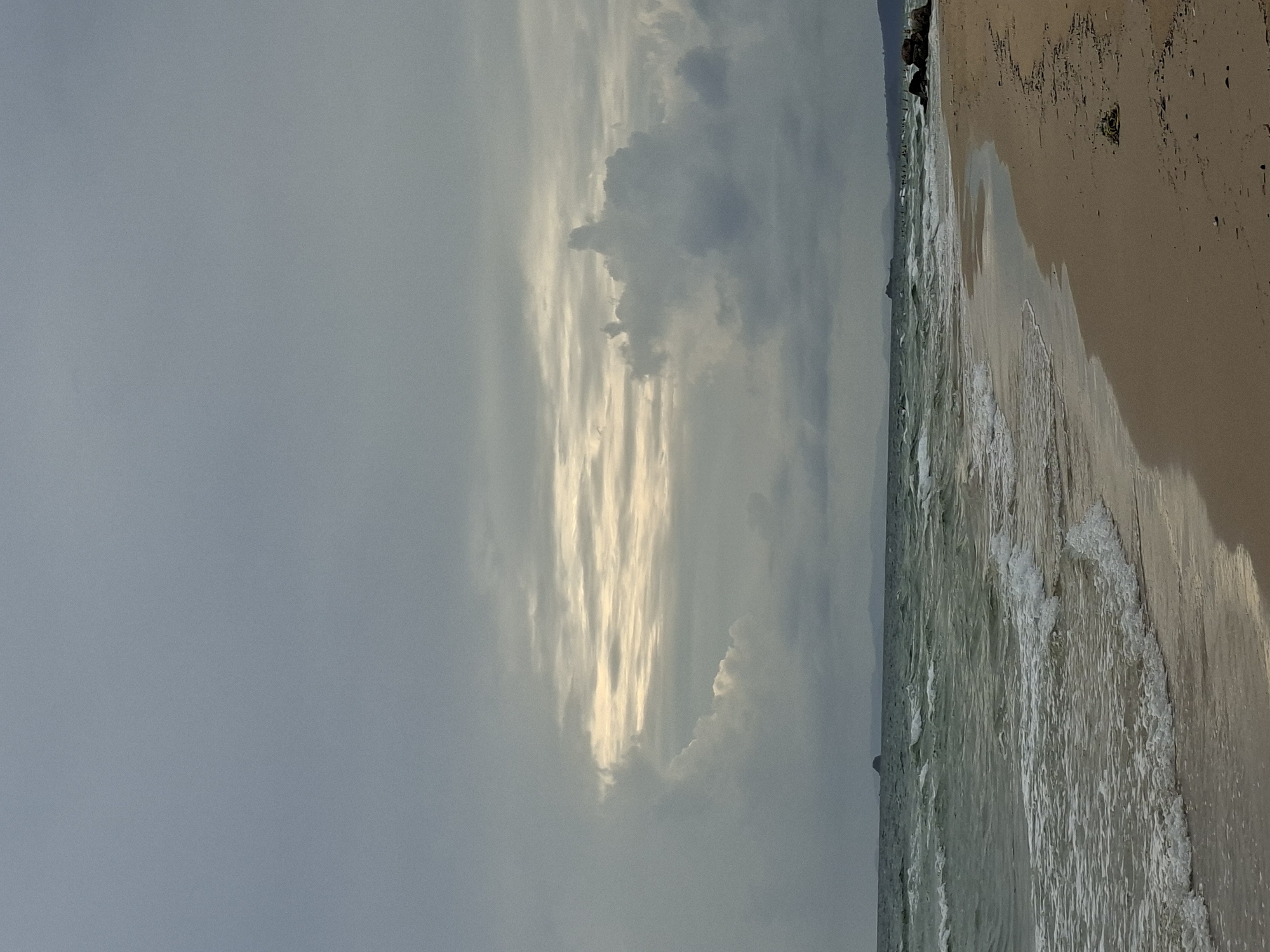 The image depicts a cloudy and somewhat overcast day at a beach in Phú Quốc, Vietnam. The foreground shows a sandy beach with a gentle slope towards the ocean. The ocean itself has moderate waves with white foam breaking near the shore. In the distance, a thin strip of land is visible on the horizon, likely the mainland or another island. The sky is dominated by large, grey clouds, with a few breaks allowing some brighter light to filter through, creating a muted, diffused illumination. There are no people or visible man-made structures in the immediate vicinity of the beach, suggesting a natural, unpopulated coastal area. The overall atmosphere is one of a calm, slightly subdued day at the seaside.