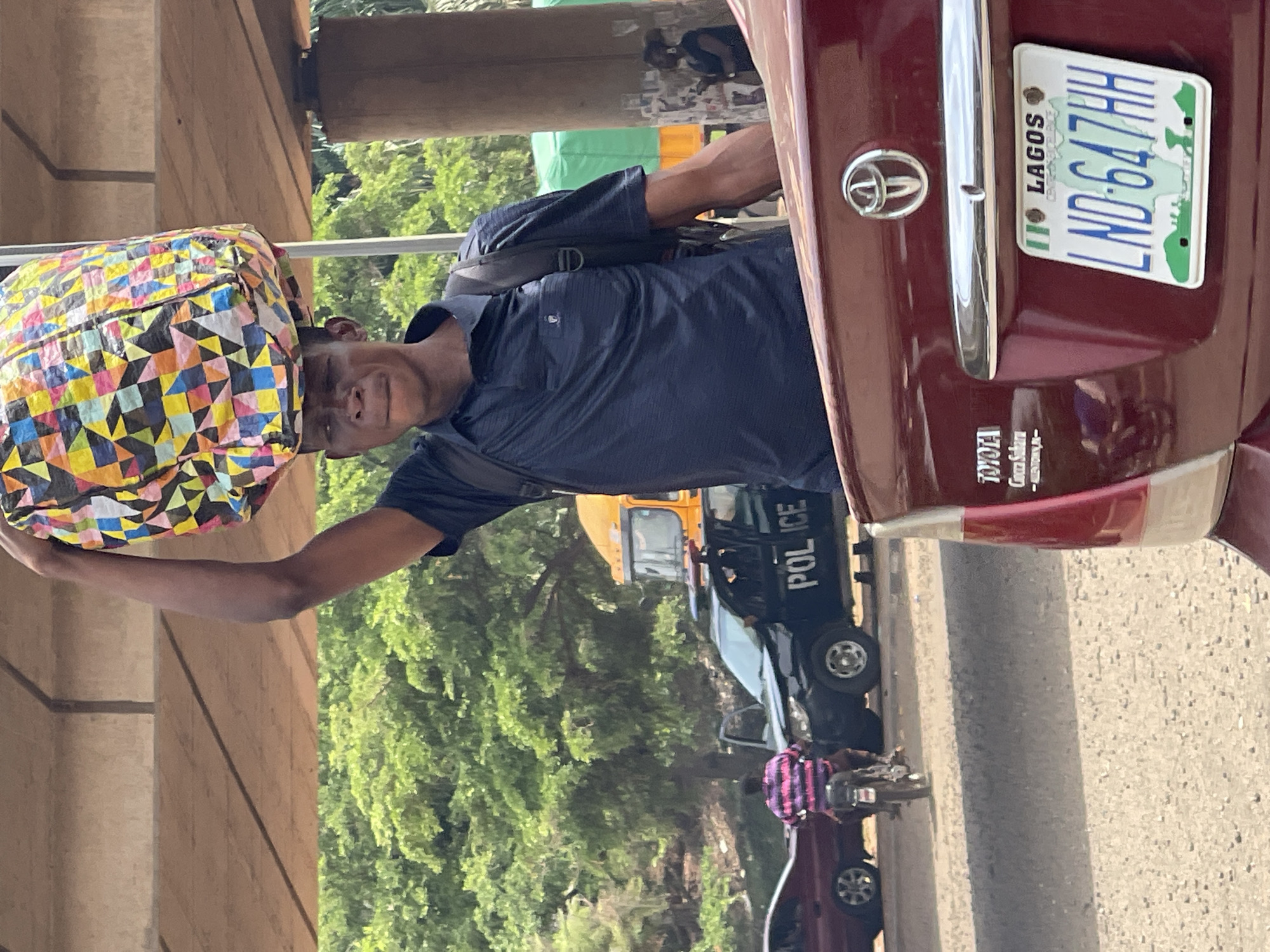 A man carries a colorful bag on his head. He is wearing a dark blue shirt and a backpack. A red Toyota car is positioned to the right. The car's license plate reads "LND-647HH" and is registered in Lagos, Nigeria. In the background, there is a police car. The scene is under a bridge, with green trees visible. The location is likely a busy street or market area in Lagos, Nigeria, given the Lagos license plate.