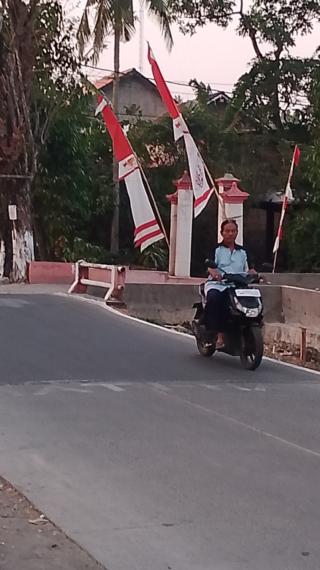 A middle-aged Indonesian man with short, dark hair and a light blue collared shirt is riding a black scooter. He is wearing dark pants and sandals. He is on a paved road that curves to the left. The road has white lane markings. Behind the road is a concrete barrier with a red and white decorative railing on top. Two white and red flags on tall poles are visible behind the barrier. The flags appear to be ceremonial. Trees and buildings are visible in the background, as well as a palm tree. The sky is overcast and light. The scene appears to be located in Karanglangu, Indonesia.