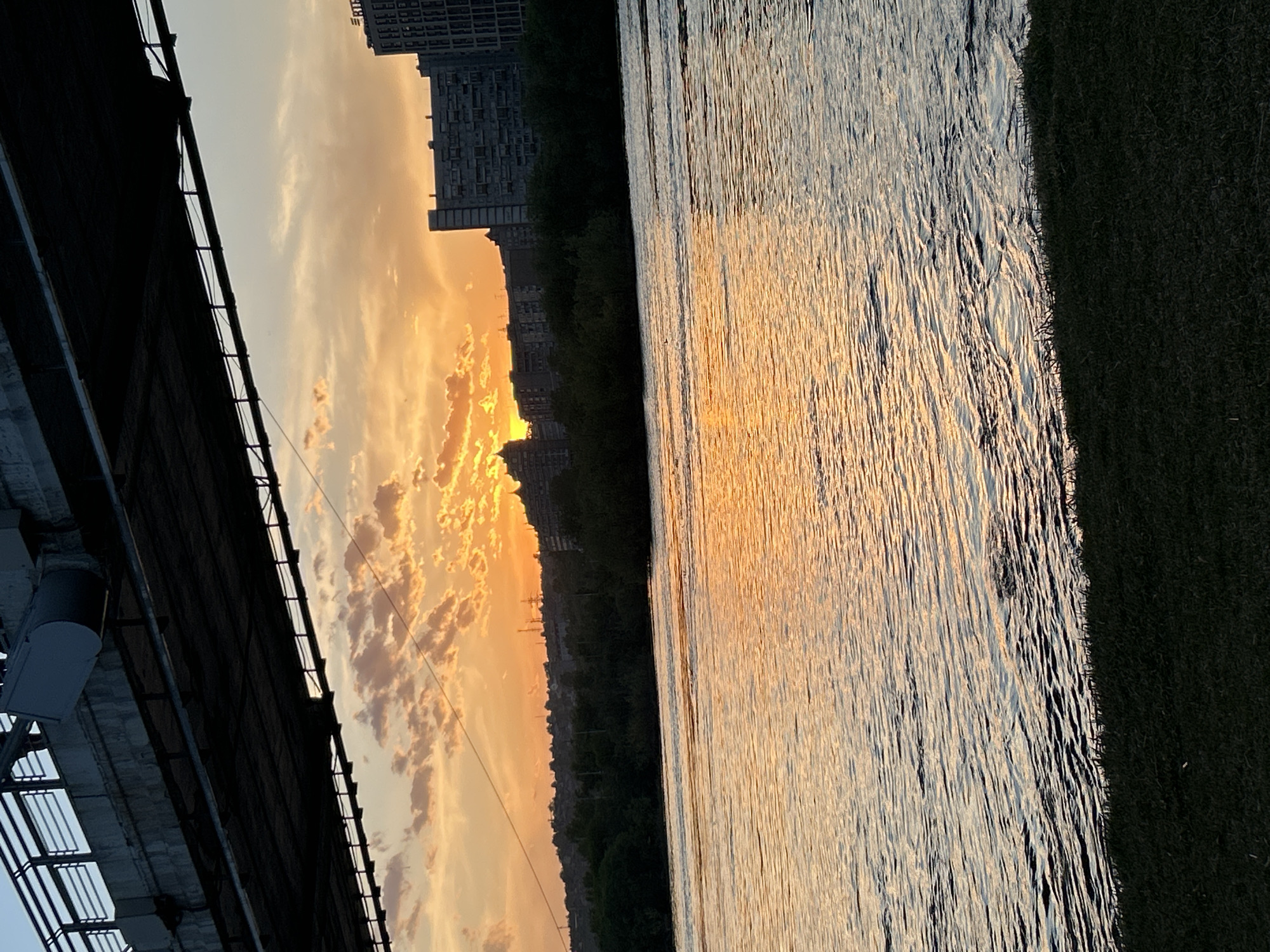 The image captures a sunset scene in Krasnoyarsk, Russia, viewed from under a bridge. The city skyline is visible against the orange and yellow sky with scattered clouds. The water surface reflects the colors of the sunset. On the right side of the image, there's a strip of grass. The underside of the bridge is in the foreground, casting shadows.