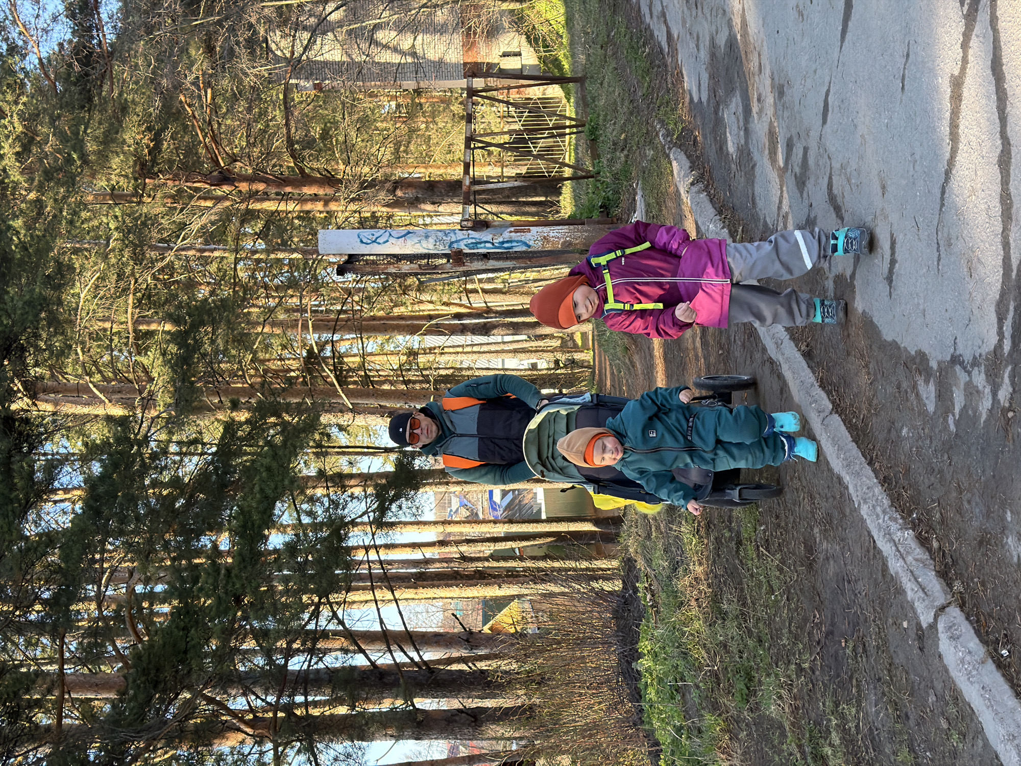 The image, oriented correctly, shows an adult male and two children on an outdoor paved path next to a wooded area. The adult, wearing a dark and orange/grey jacket, sunglasses, and a black cap, is pushing a black and green stroller or wagon. A young child, dressed in a dark green jumpsuit with a small white square logo and an orange-brown hat, sits in the stroller, looking towards the camera.
To the adult's right, standing on the paved path, is a second child. This child wears a purple jacket with lime green straps, grey pants with a reflective strip, an orange-brown hat, and dark shoes with turquoise details. This child also looks towards the camera.
On the left side of the scene, a dense collection of tall, slender pine trees creates a backdrop, with sunlight filtering through them and casting long shadows across the ground and path. A long, white concrete or metal barrier with abstract blue graffiti is situated between the trees and the path. Behind this barrier, a rusty, dilapidated metal structure is visible, and further in the background, a building can be vaguely discerned through the trees.
The paved path itself has several wet patches or puddles. The clear lighting suggests a sunny day, likely late afternoon or early morning. The scene depicts a family outing, with the group appearing to be strolling or walking. No significant discernible text is visible other than a small logo on the child's jumpsuit.