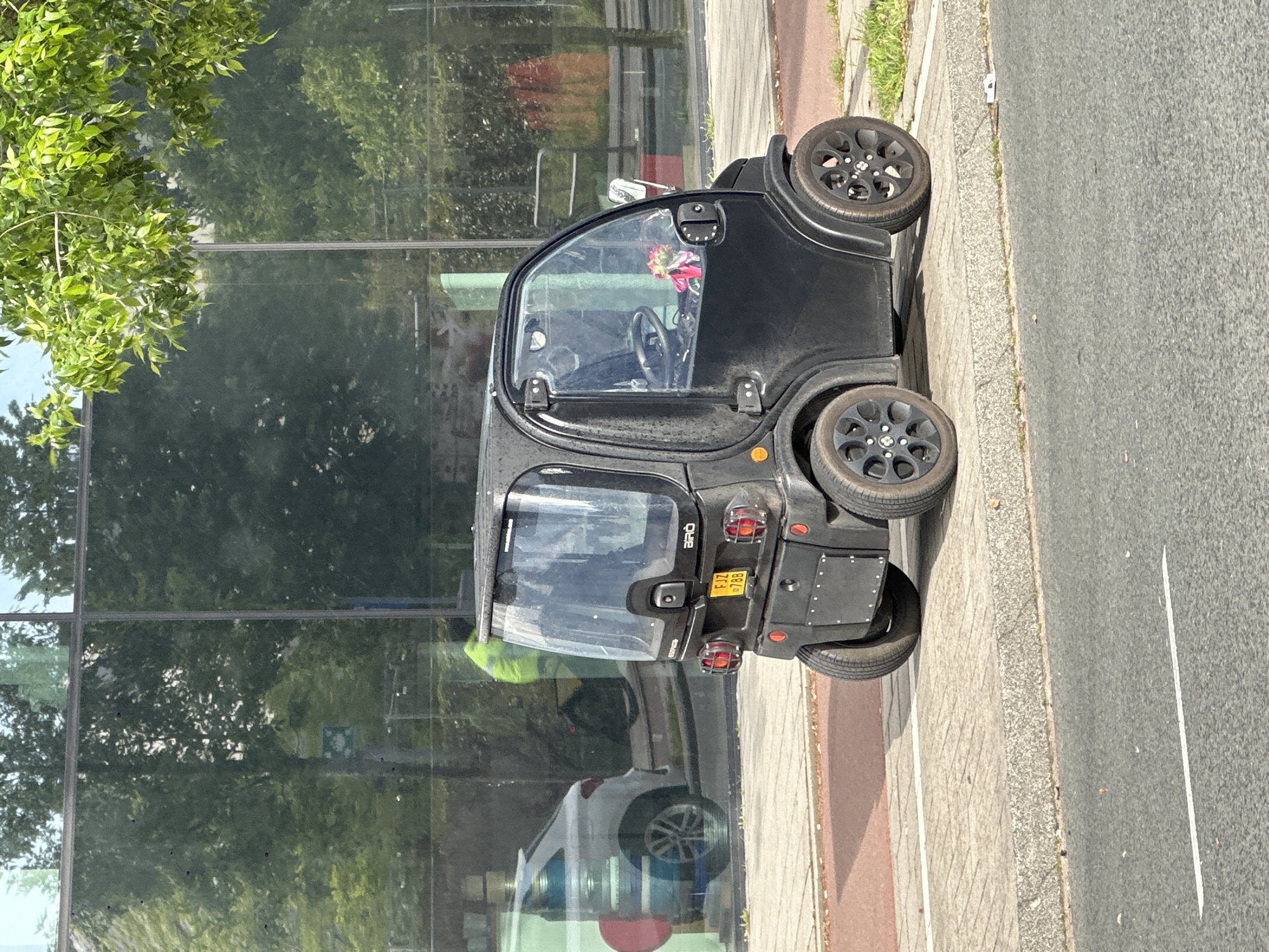 The image depicts a black AIXAM e-City electric quadricycle parked on a sidewalk adjacent to a road in Amsterdam, Netherlands. The vehicle has a Dutch license plate reading "FJ-17-788". In the driver's seat, a bouquet of flowers is visible. The surrounding environment reflects in the glass facade of a building, showing trees and parts of vehicles.