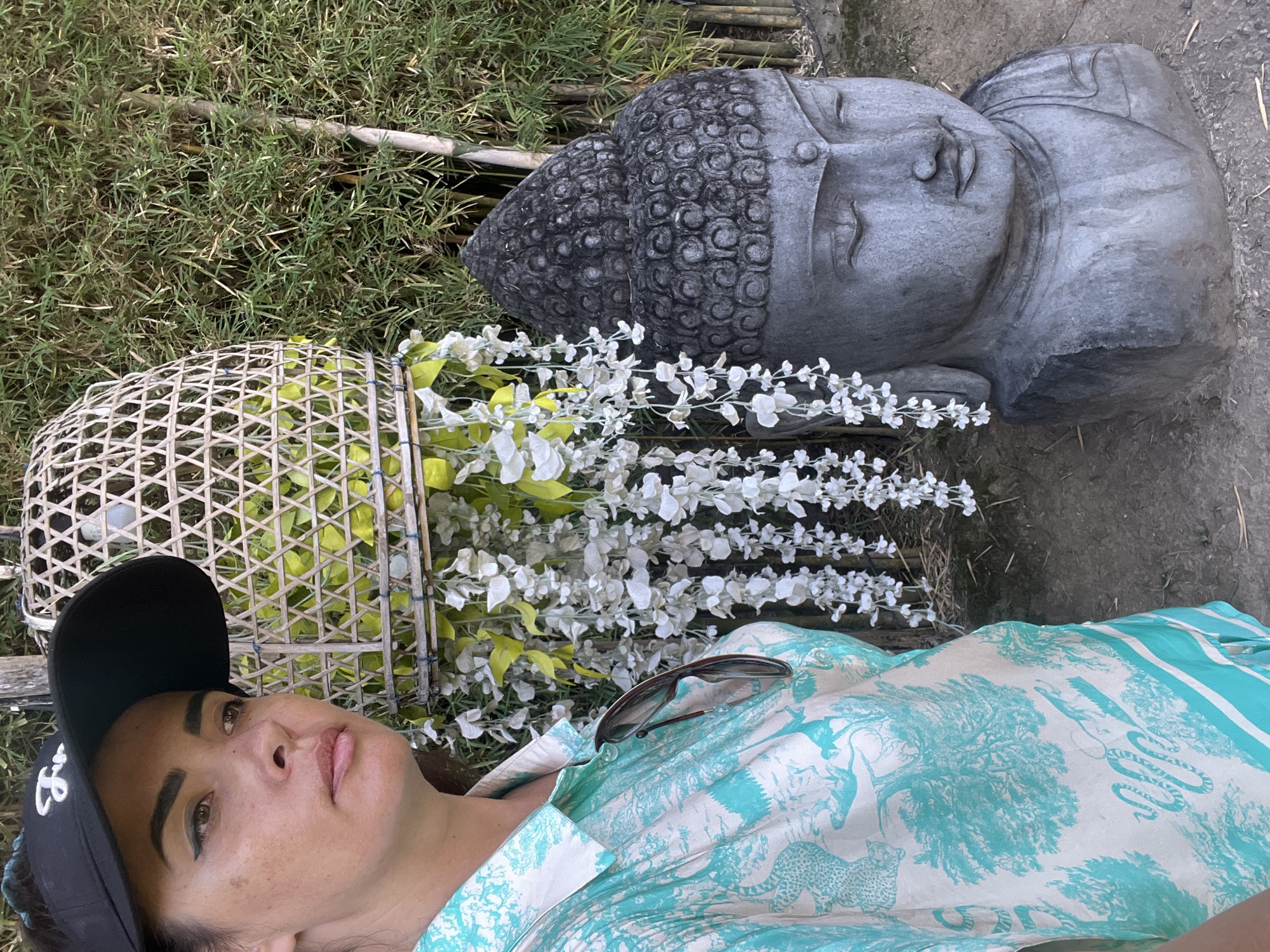 The image shows a woman wearing a black visor and a turquoise and white patterned shirt. She is standing next to a stone statue of Buddha's head. Behind them is a bamboo forest and a decorative woven basket with white flowers hanging from it. The scene appears to be an outdoor setting, possibly a garden or temple in Denpasar, Indonesia. The woman is looking to her left, with sunglasses hanging from her shirt. The Buddha statue is facing forward, with its eyes closed in a meditative pose. The bamboo stalks are tall and dense, creating a natural backdrop. The woven basket is intricate in design, adding a cultural element to the scene.