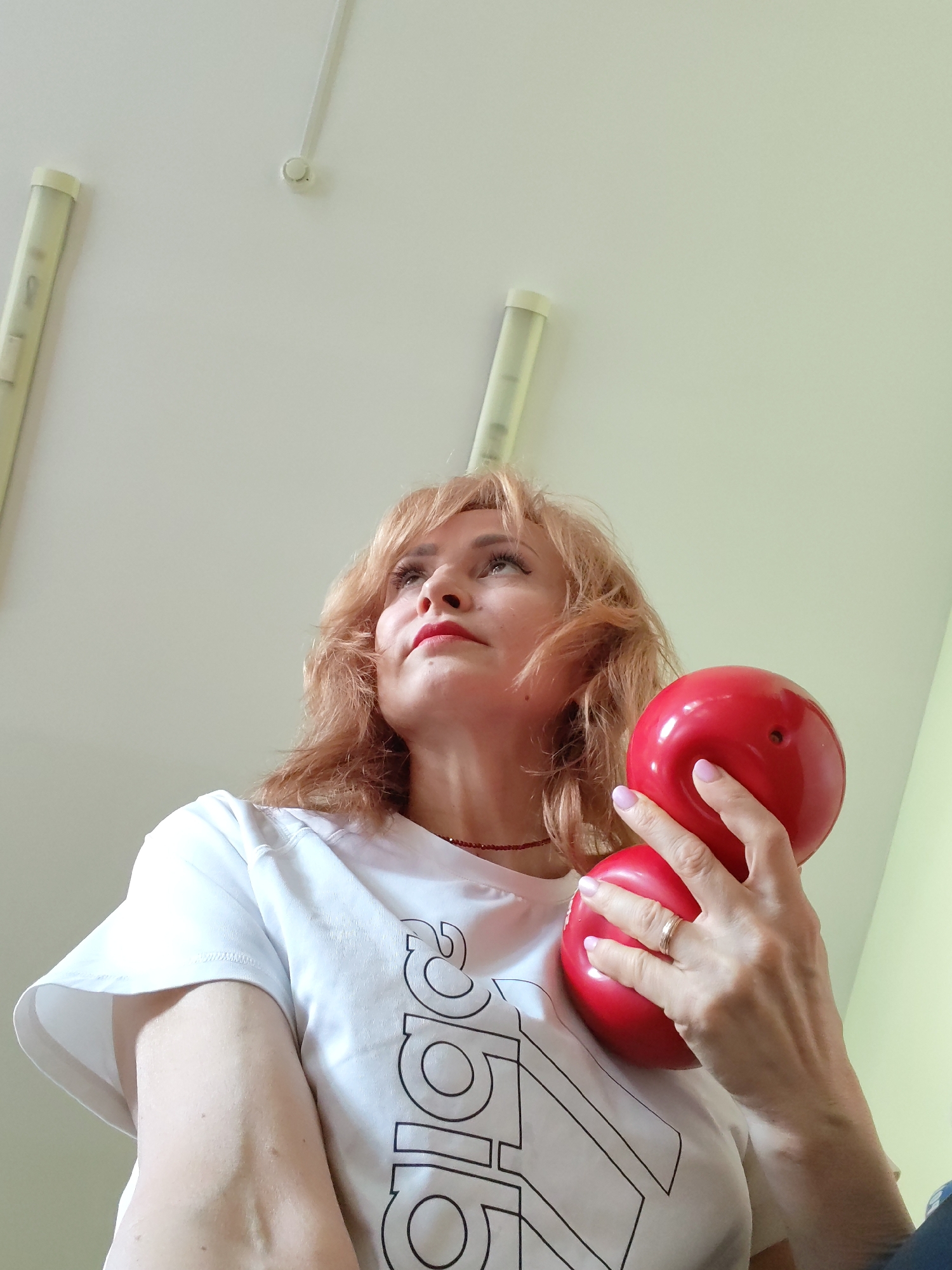 A fair-skinned woman with short, reddish-blonde hair, wearing a white t-shirt with black lettering, is holding a red hourglass-shaped object. She is looking upwards and slightly to her left. She is wearing a red beaded necklace and a gold ring on her left ring finger. The background is a white ceiling with two light fixtures, and a pale green wall. The location is Chaikovsky, Russia, as the information was provided with the context of the image.