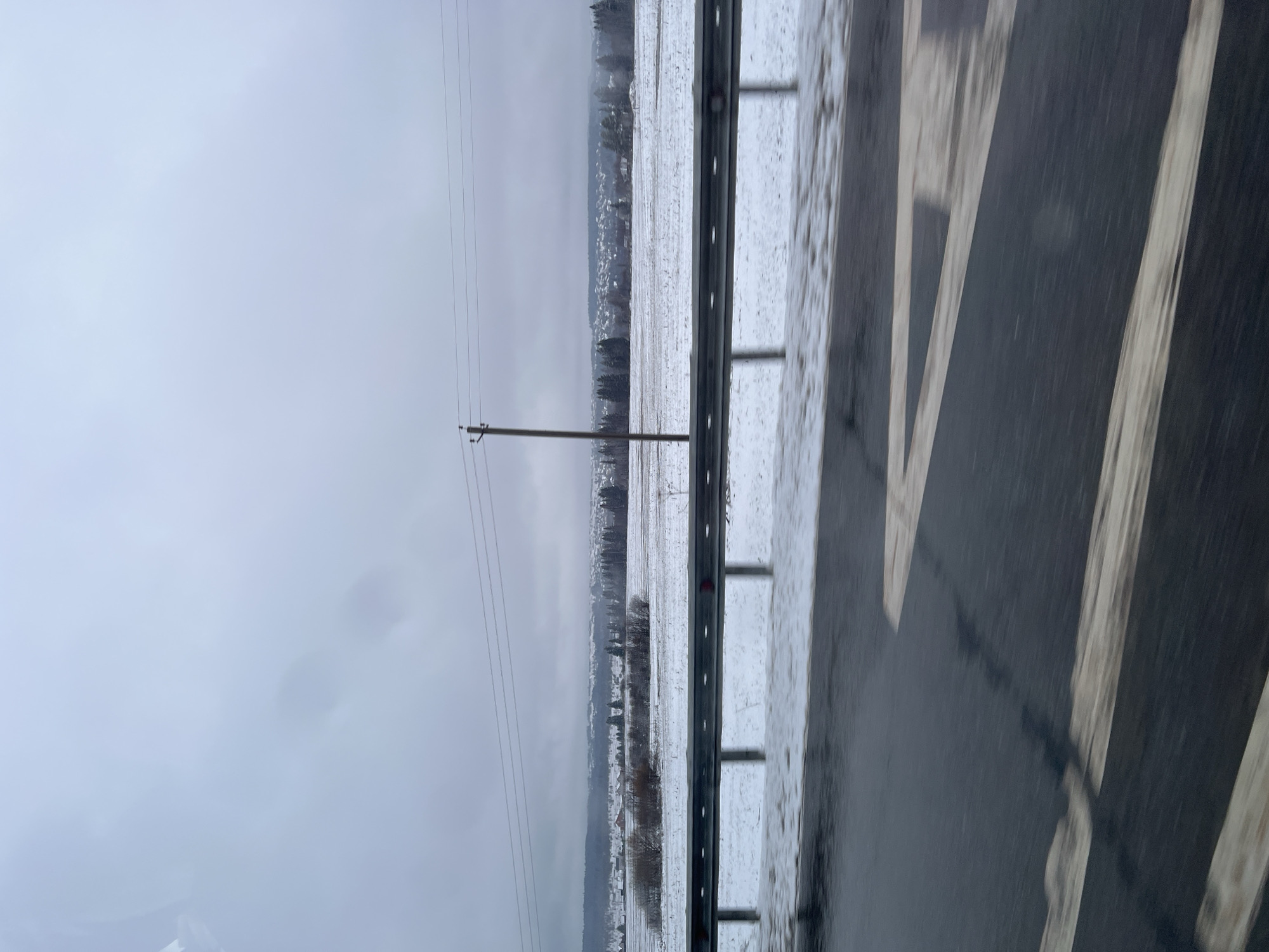 The image shows a snowy landscape viewed from a moving vehicle, likely on a highway. On the right side of the frame is a blurred view of the wet asphalt road with painted white lines. A guardrail runs along the roadside. Beyond the guardrail is a snow-covered field leading to a distant treeline and a faint, indistinct settlement on the horizon, possibly the outskirts of a town. A utility pole with power lines is visible in the mid-ground. The sky is overcast and gray. The overall scene suggests a rural area in winter. The location of Kosoturikha, Russia is implied and does not show any specific details in the image to verify it.
