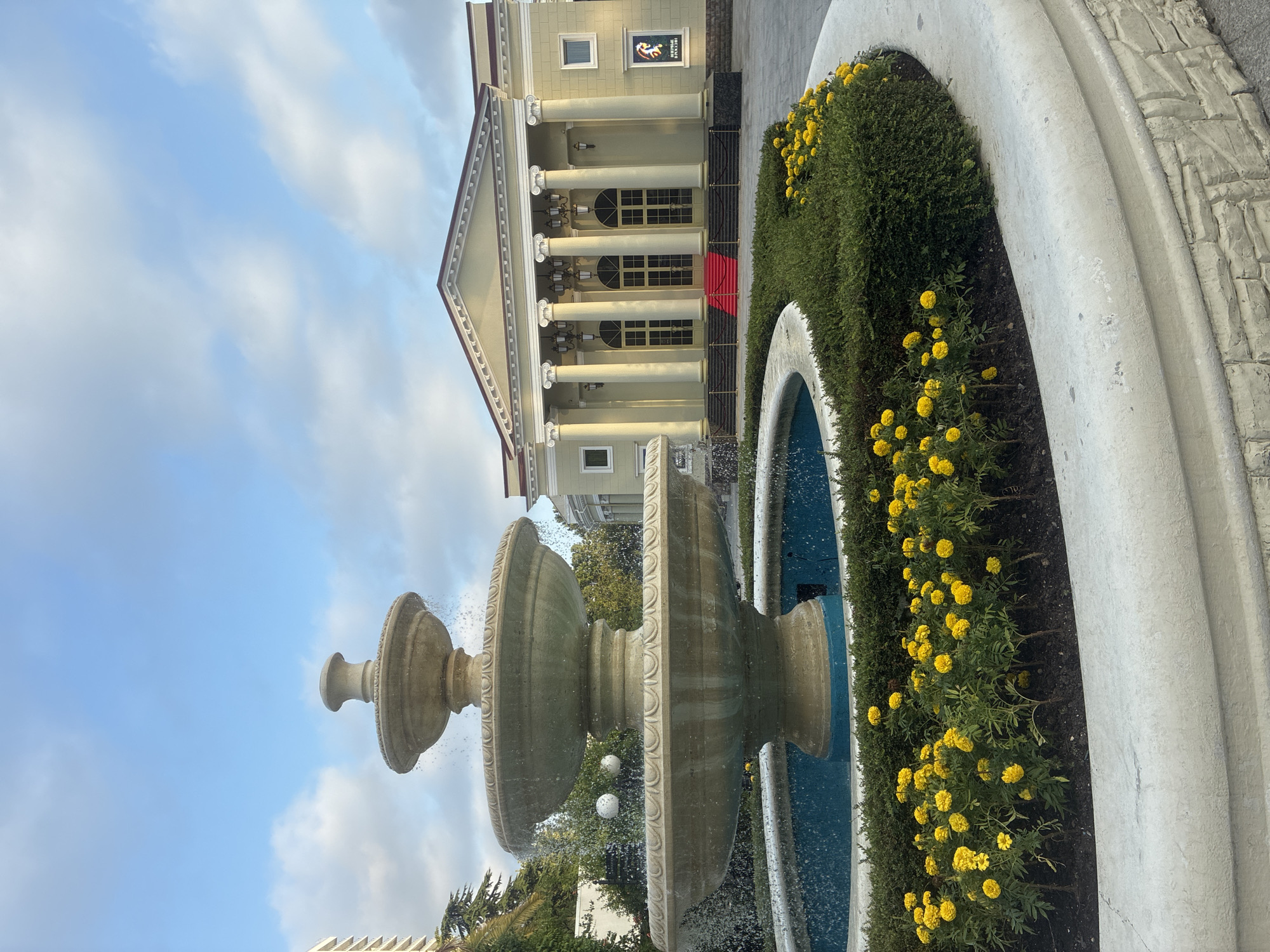 The image shows a multi-tiered fountain in the foreground, with water spraying from its upper levels into a blue basin. The fountain is surrounded by a curved white concrete planter filled with manicured green shrubs and numerous small yellow flowers, likely marigolds. Behind the fountain, a pale yellow building with a classical facade stands prominently. This building features a portico with tall white columns, a triangular pediment, and large arched windows. A red carpet leads up to the entrance. The sky above is a mixture of blue and white clouds. A poster featuring a horse is visible on the right side of the building's exterior. To the left of the fountain, a portion of a modern building with balconies can be seen, along with some trees. This scene is located at the Sochi Dendrarium in Sochi, Russia. There are no people visible in the image.