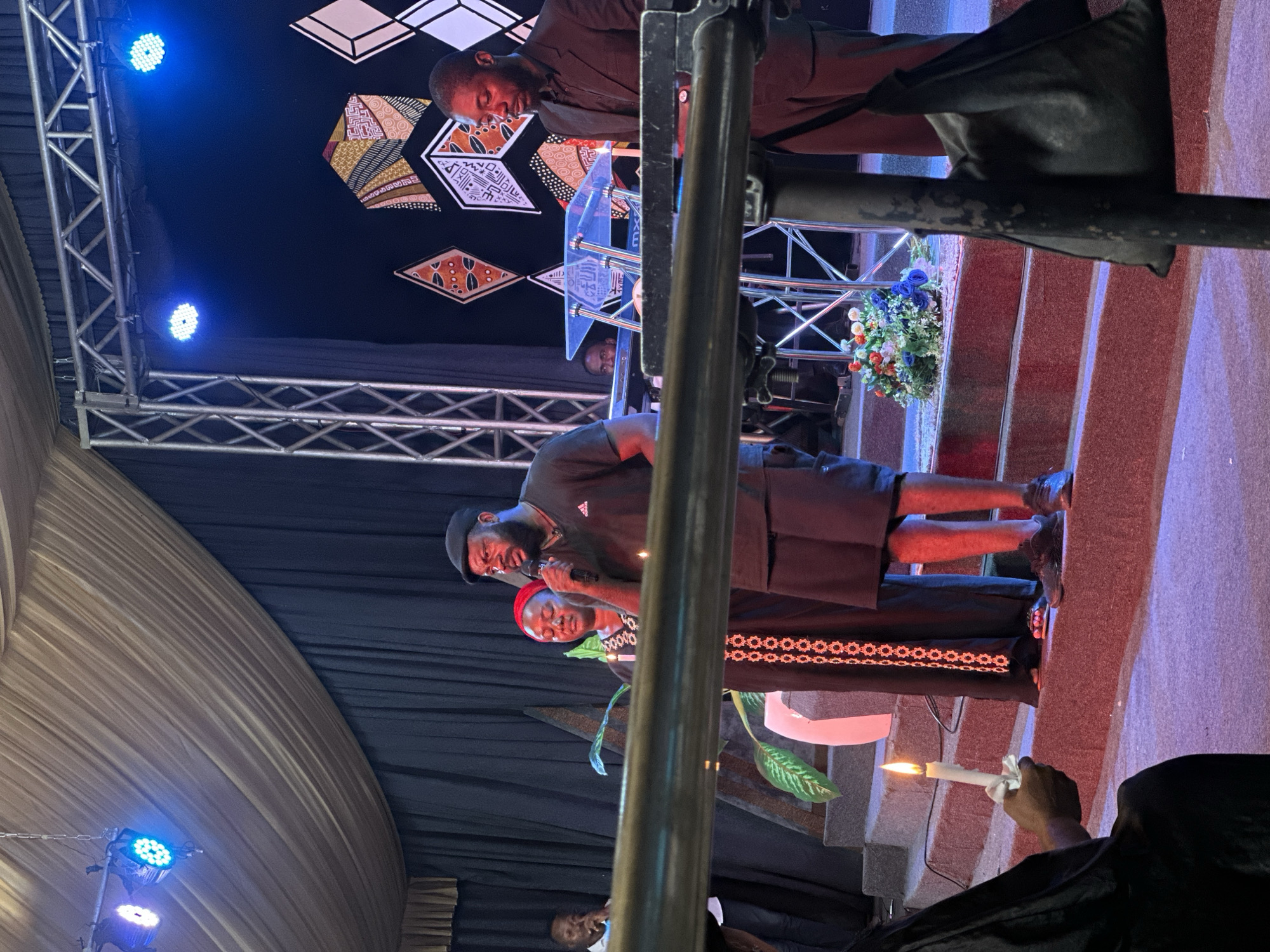 The image depicts a gathering in Ashipa, Nigeria. The scene appears to be an indoor event, possibly a service or ceremony, taking place on a stage.
In the foreground, on the left, a person in black clothing is holding a lit candle. To the right of this person are three individuals standing on elevated steps. The central figure is a man wearing a black t-shirt and shorts, holding a microphone and speaking. To his left is a woman wearing a traditional black dress with red and white embroidery, and a red headscarf. She is also holding a lit candle. To the right of the man with the microphone, a portion of another man in a black suit is visible.
Behind the main group, there's a stage with black curtains. A floral arrangement sits on the steps. The background features a decorative wall with geometric patterns in shades of brown, yellow, and white. Stage lighting, including blue spotlights, illuminates the area. A metal truss structure is visible above the stage. On the right side of the stage, there's a transparent podium.
The overall impression is one of a formal gathering, with elements of reverence suggested by the candles and the solemn expressions of some individuals.