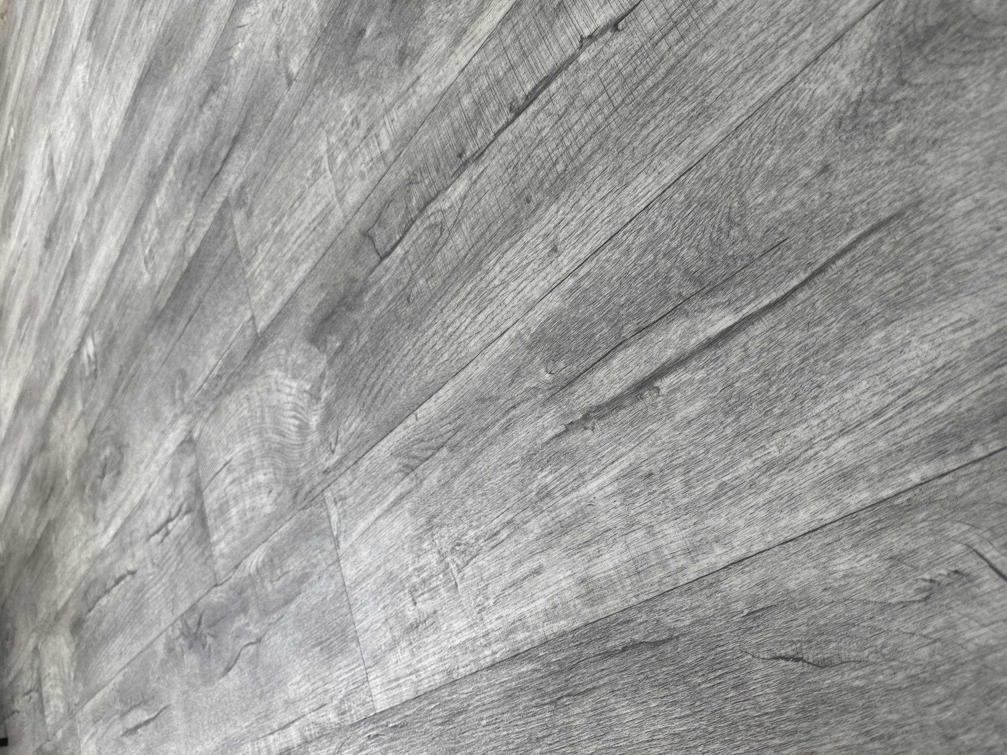 This is a close-up, angled shot of a gray wood-look floor. The planks are laid parallel to each other and run diagonally from the bottom left to the top right of the frame. The texture of the floor suggests it is laminate or vinyl flooring, mimicking the grain and imperfections of natural wood. The lighting creates subtle shadows along the seams between the planks, highlighting the horizontal grain pattern. There are no people, objects, or visible environmental features other than the flooring itself. The location within Tabuny, Russia, is not discernible from the image alone.