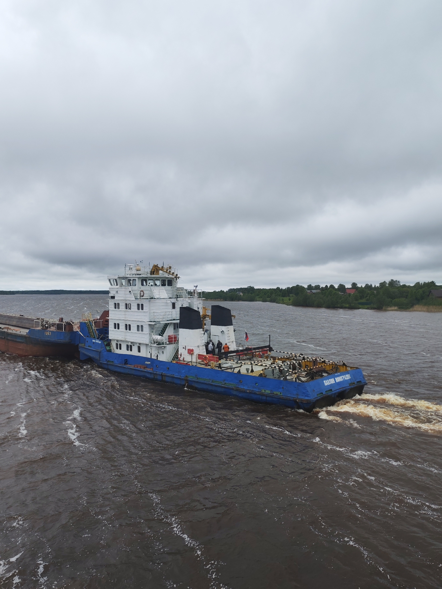A blue and white tugboat, identified as "PAVEL VINOGRADOV" in Cyrillic script, is moving across the water, leaving a wake. A barge is attached to its side. The location appears to be near Анхимово, Russia, judging by the river landscape and construction of the tugboat. The sky is overcast. Trees are visible on the shoreline in the background. There are several people on the boat.