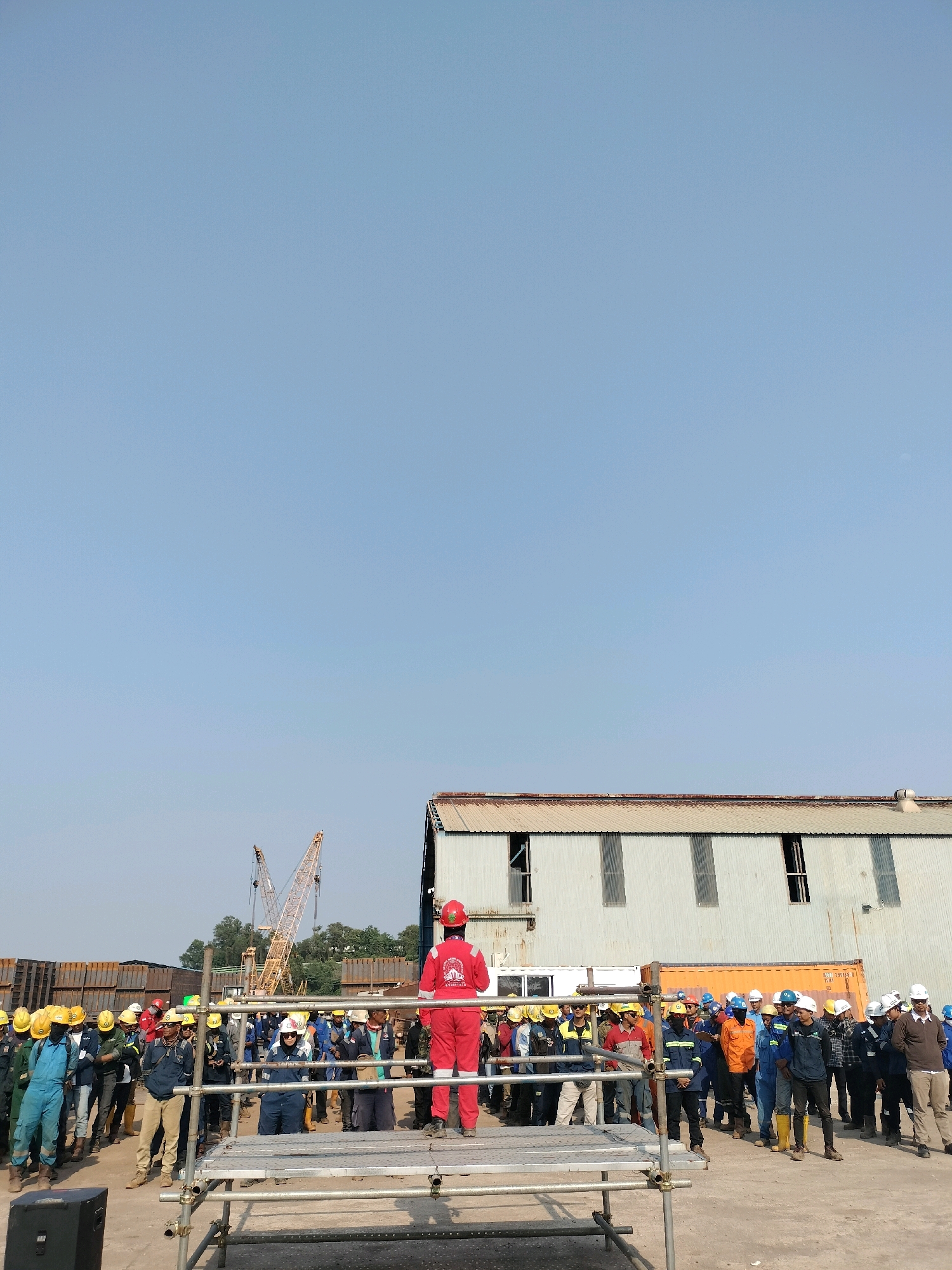 An outdoor daytime scene in Batam, Indonesia, shows a group of workers gathered in what appears to be an industrial or construction site. In the foreground, a person in a red jumpsuit and red hard hat stands on a scaffolding, facing away from the camera and addressing the assembled group. The crowd, composed of approximately 30-40 individuals, is dressed in various work attire, predominantly including hard hats in yellow, white, and red, and some are wearing safety vests over their clothes. Many of the individuals are looking towards the person on the scaffolding, suggesting a briefing or assembly.
The background features industrial elements such as shipping containers, cranes, and large building structures with corrugated metal exteriors. The overall environment is flat and dusty, with a clear blue sky overhead. The presence of industrial equipment and the attire of the people strongly indicate an operational industrial facility or construction zone within Batam. The lighting suggests it is daytime with bright sunlight.