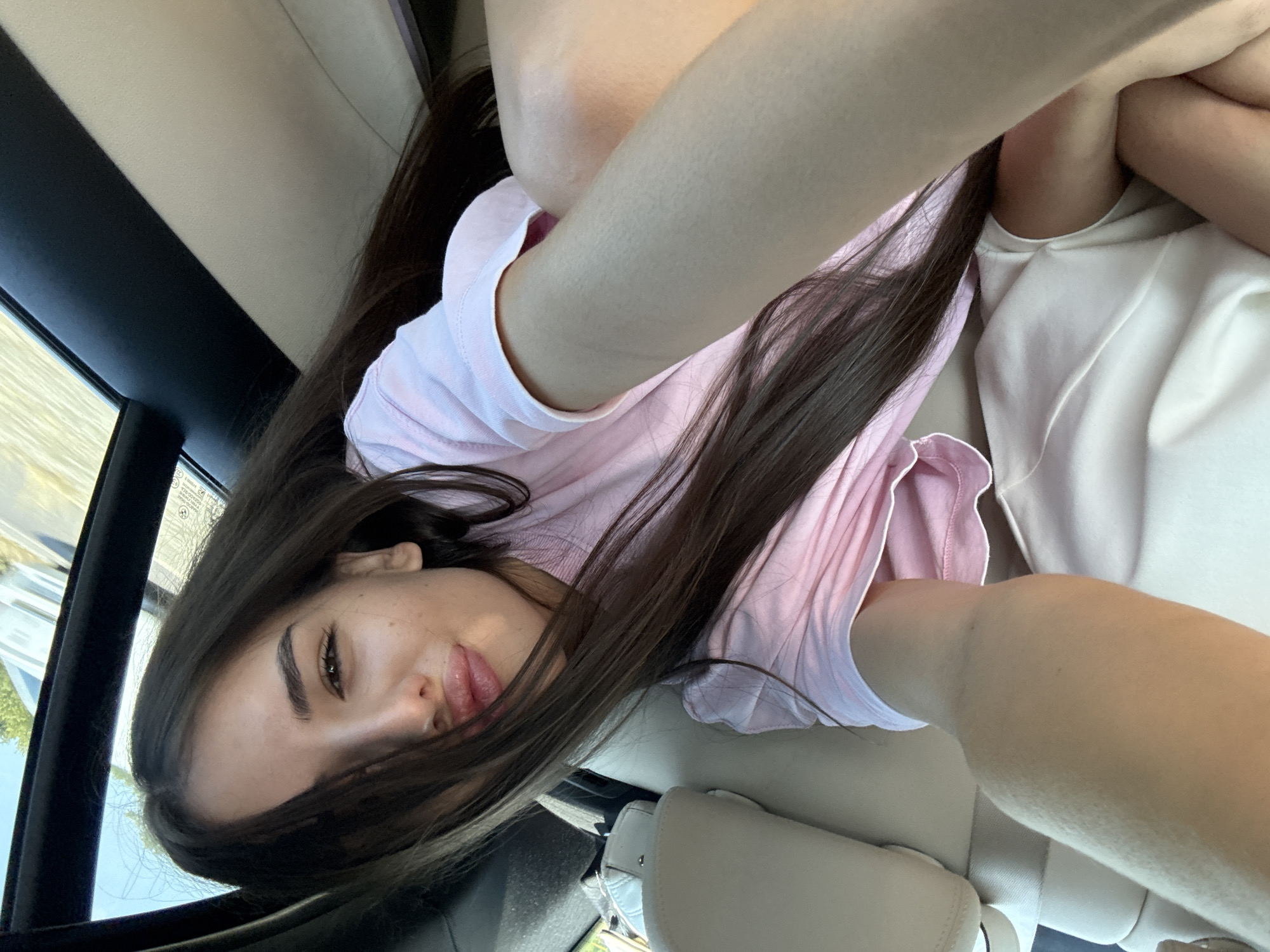 A young woman with long dark hair and fair skin is pictured in the backseat of a car, taking a selfie. She is wearing a pink t-shirt and a white garment that appears to be part of her clothing or a blanket. Her right arm is extended towards the camera, and her face is partially obscured by her hair. She is looking towards the camera with her lips pursed. The interior of the car is visible, with a light-colored seat and a black door panel. The windows show a blurred outdoor scene of trees and a road, suggesting the car is in motion. The scene does not contain any elements specific to Ucea de Jos, Romania, beyond the possibility of it being a general road or travel location.