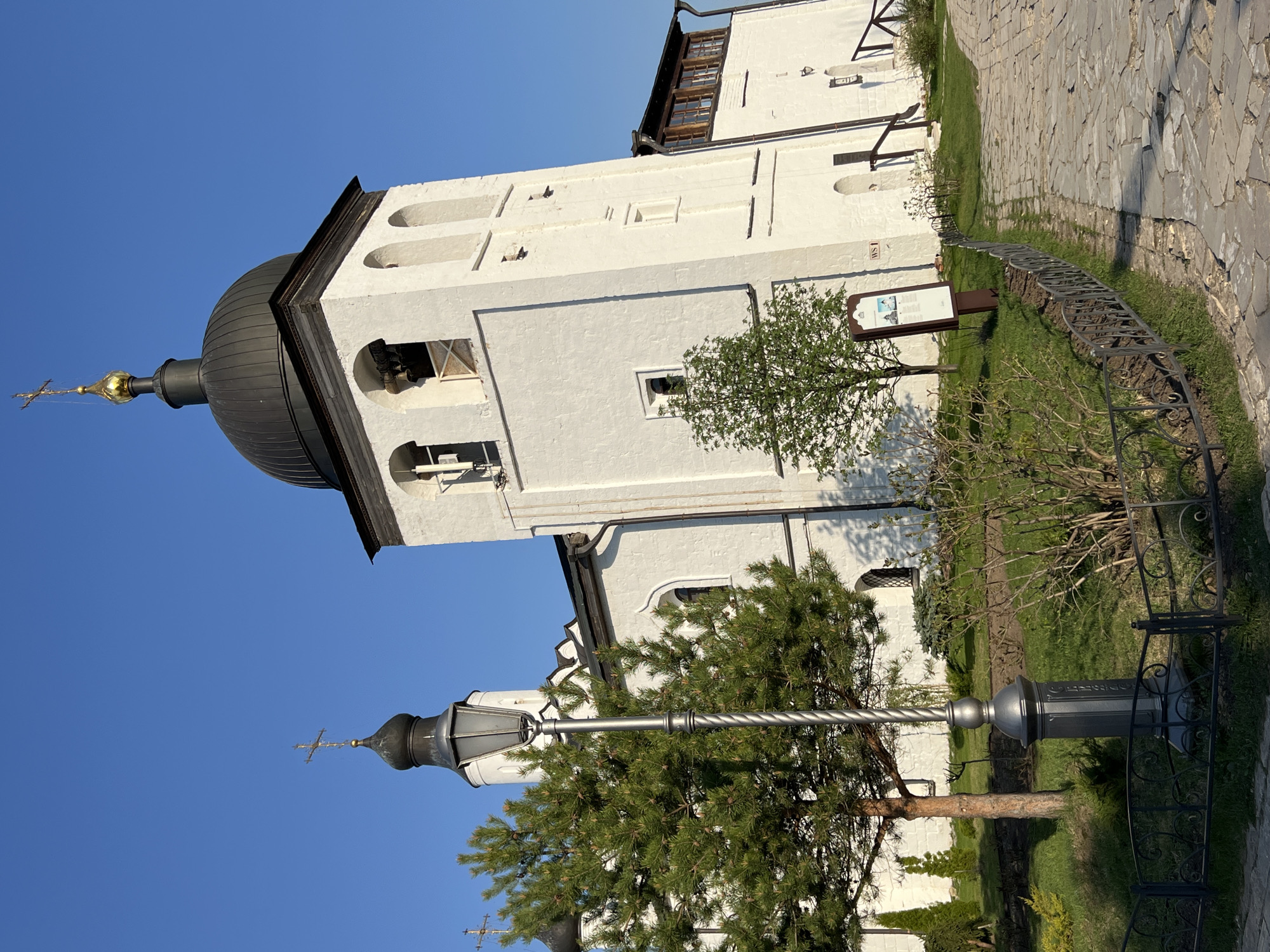 The image captures a vertical view of the Sviyazhsk Holy Assumption Monastery in Sviyazhsk, Russia. The building complex is predominantly white with dark grey or black domes and accents. The architectural style includes arched windows and classical Russian Orthodox church elements.
The foreground features landscaping, including trees and a metal fence separating a stone pathway from the grass and foliage. A street lamp is visible, partially obscured by a pine tree. The sky is a clear, bright blue. There are no people visible in the frame.