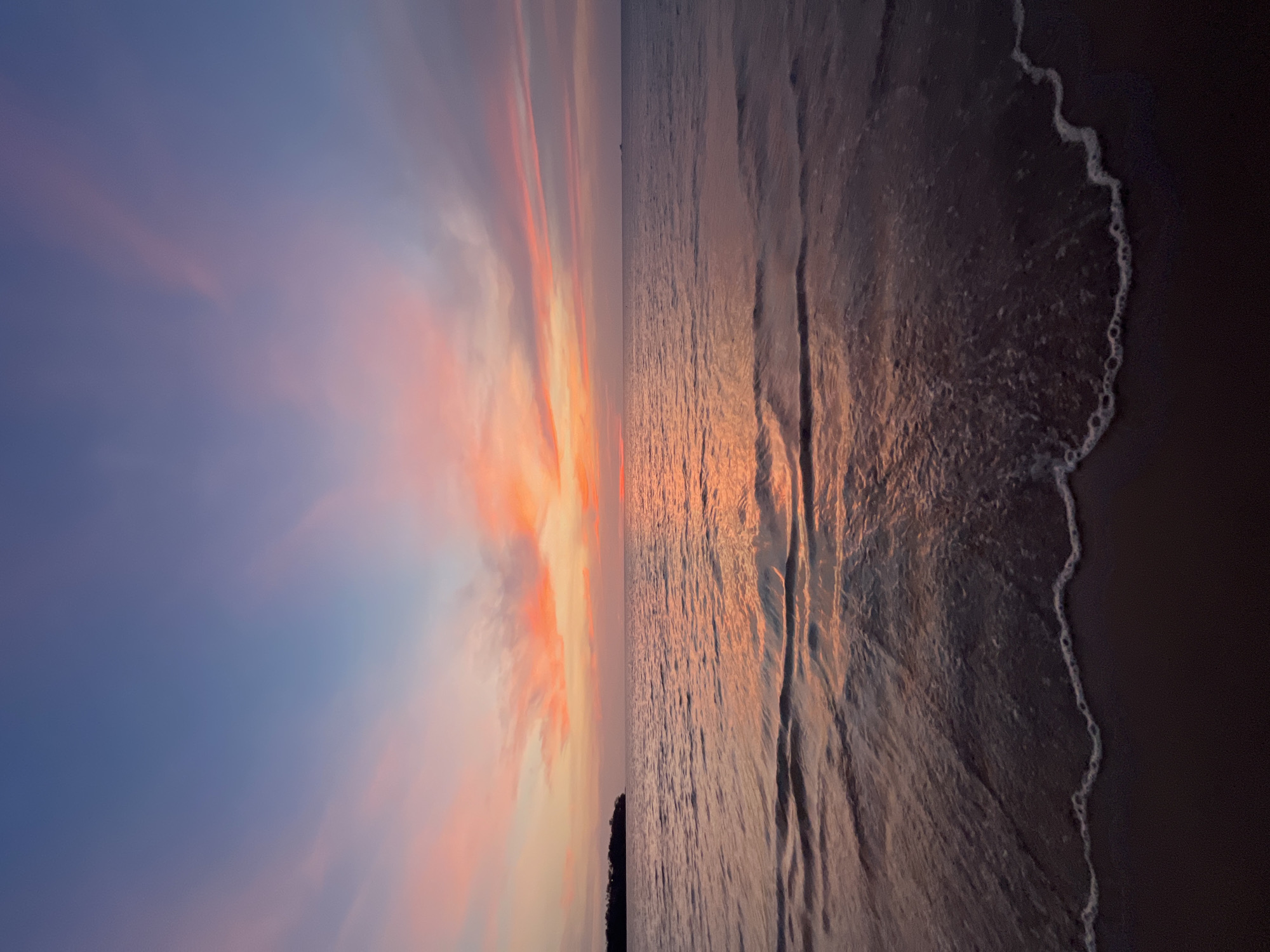 The image depicts a sunset scene at an unknown beach in the city of Unknown, Unknown. The sky displays a gradient of colors, transitioning from blue to orange and pink hues, suggestive of sunrise or sunset. The beach is a dark color, with waves gently lapping the shoreline. The water reflects the colors of the sky, creating a mirror-like effect. A sliver of trees can be seen in the bottom left corner, providing a sense of depth to the scene. There are no people or other identifiable objects present.