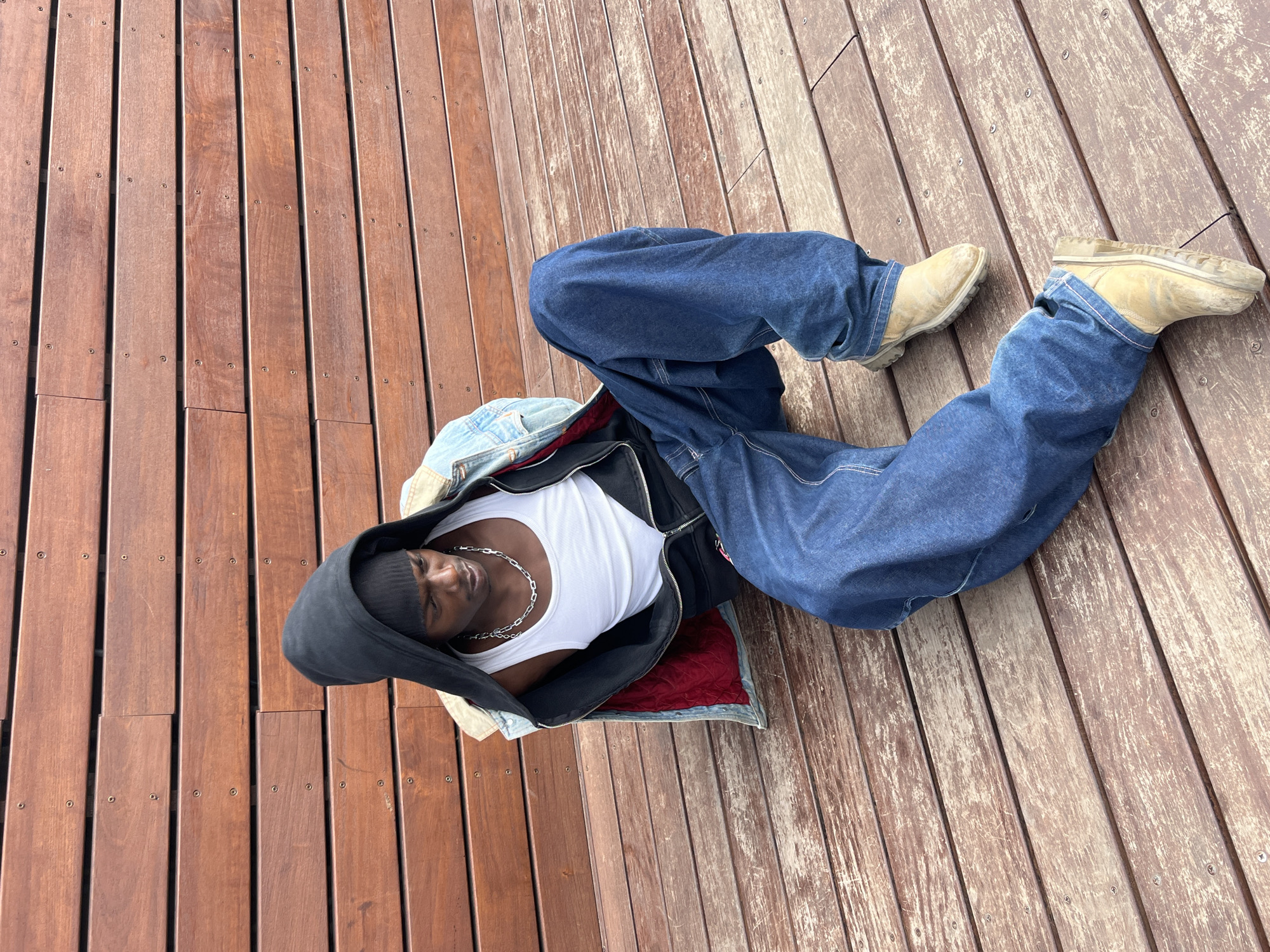 A young Black man is lying on a wooden deck. He is wearing a black hoodie, a white tank top, a silver chain, a black jacket, a denim jacket, baggy blue jeans, and tan work boots. The wooden deck is made of parallel planks of wood, and it appears to be partially in the sun and partially in the shade. The location cannot be determined to be Barcelona, Spain from the image.