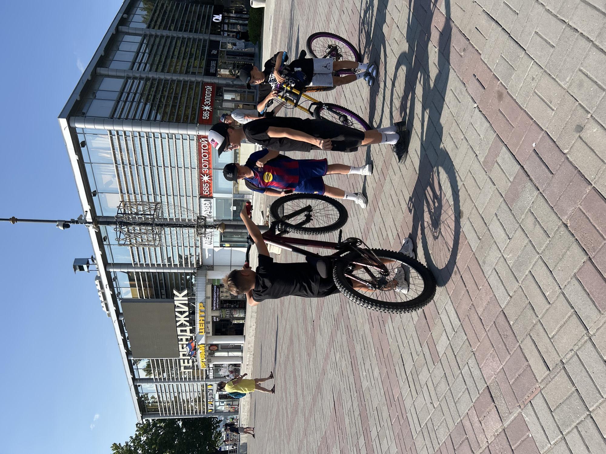 A group of four young boys are gathered on a paved plaza in front of a modern building with large glass windows. The building has a prominent sign that reads "ГЕЛЕНДЖИК" (Gelendzhik) in large white letters, and below it, "ОРГОВЫЙ ЦЕНТР" (Trade Center). The plaza is paved with rectangular bricks in a pattern of red and grey. The boys are all holding or standing near bicycles. One boy in the foreground on the left is wearing a black t-shirt and shorts and is holding a maroon bicycle upright by its handlebars, with the front wheel lifted off the ground. To his right, two other boys are standing with bicycles. One boy is wearing a blue and red striped soccer jersey with blue shorts. The other boy is wearing a black t-shirt and shorts, and a black baseball cap. To the far right, a fourth boy is standing with a yellow and black bicycle. In the background, other people are walking on the plaza, and there are shops visible within the trade center building. The sky is clear and blue, indicating a sunny day. The scene appears to be in the city of Gelendzhik, Russia, as indicated by the building's signage.