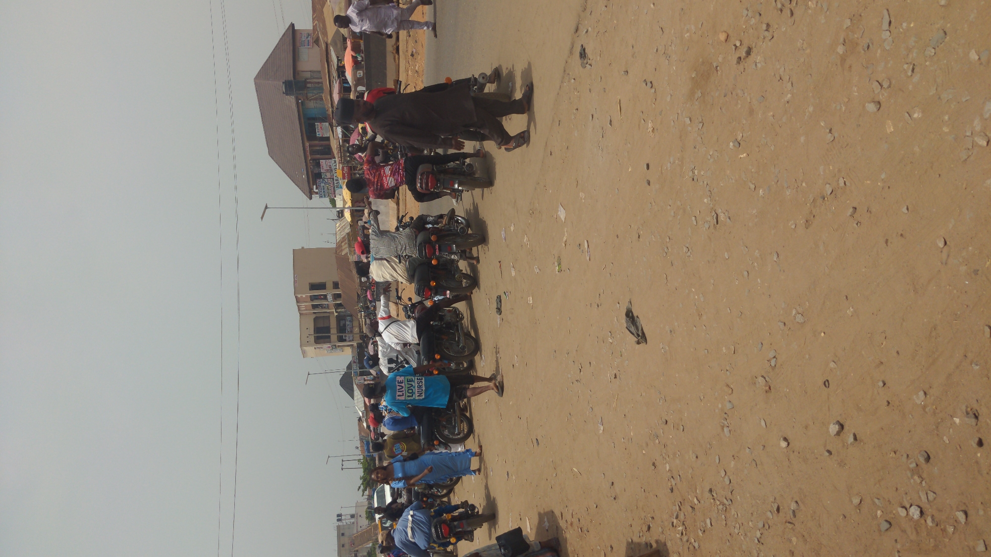 This image captures a busy daytime street scene in what appears to be a town or city in Ado, Nigeria. The dominant feature is a wide, unpaved dirt road, light brown in color, with scattered small rocks and debris.
Numerous people are present, engaged in typical street activities. Many individuals, mostly men, are seen standing or sitting on motorcycles, suggesting a common form of transport like motorcycle taxis (okadas) or a gathering point for riders. Other people are seen walking along the roadside. The overall impression is one of everyday hustle and bustle.
Along the left side of the road, various buildings are visible. These structures appear to be a mix of residential and commercial establishments, with some showing signs of shops or businesses. One building in the mid-ground has a distinct dark brown roof. Overhead electrical or communication wires stretch across the top portion of the frame against a bright, clear sky, indicating sunny weather.
Notable details include the prevalence of motorcycles, the unpaved nature of the road, and the mix of clothing styles worn by the people.
Visible text includes:
* On a bright blue t-shirt worn by a person near the center-left, the words "LIVE LOVE NURSE" are clearly legible in white capital letters.
* On a sign on a building in the mid-background, partial text "HARO. BAM" can be seen.