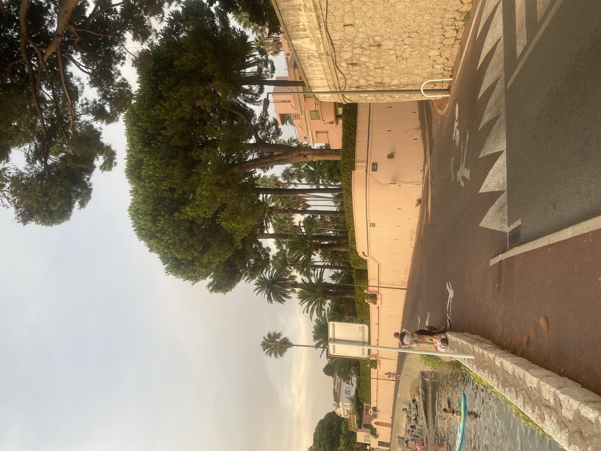 The image shows a scenic view of the coastline in Antibes, France. A paved road curves along a stone embankment bordering the Mediterranean Sea. On the left, a rocky beach is dotted with people relaxing and one person paddleboarding. The road is marked with pedestrian and cyclist symbols, indicating a shared pathway. A pale pink building with arched doorways is visible behind a low wall adorned with palm trees. Further up the hill, a larger pink building with a curved facade stands amongst lush greenery. Tall pine trees and various palm trees frame the scene, with the sky overcast. The overall impression is one of a tranquil seaside setting in the late afternoon light.