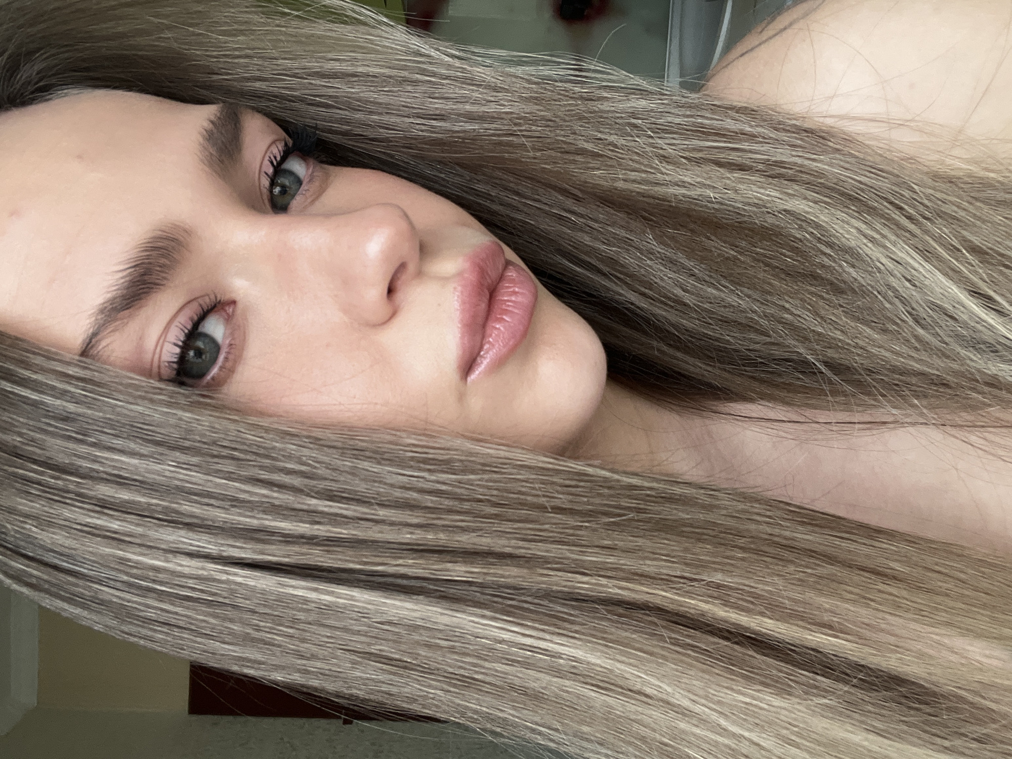 A close-up, vertical portrait captures a young adult female from the chest up, positioned indoors in the city of Unknown, Unknown. The subject is centered, looking directly towards the camera with a slight head tilt. She possesses long, straight, multi-tonal blonde and light brown hair that frames her face and extends downwards, obscuring portions of the background. Her eyes are blue-green/hazel, with prominent eyelashes showing evidence of mascara or enhancement. Eyebrows are dark and defined. Her nose is straight. The subject's lips are full, slightly parted, and coated with a glossy pink product. Skin tone is fair. A bare shoulder is visible on the right side of the frame. The background, visible primarily behind her left shoulder, is out of focus and consists of a light beige or cream-colored wall section, with a partial view of a vertical reddish-brown object, likely a door frame or piece of furniture, further in the background.