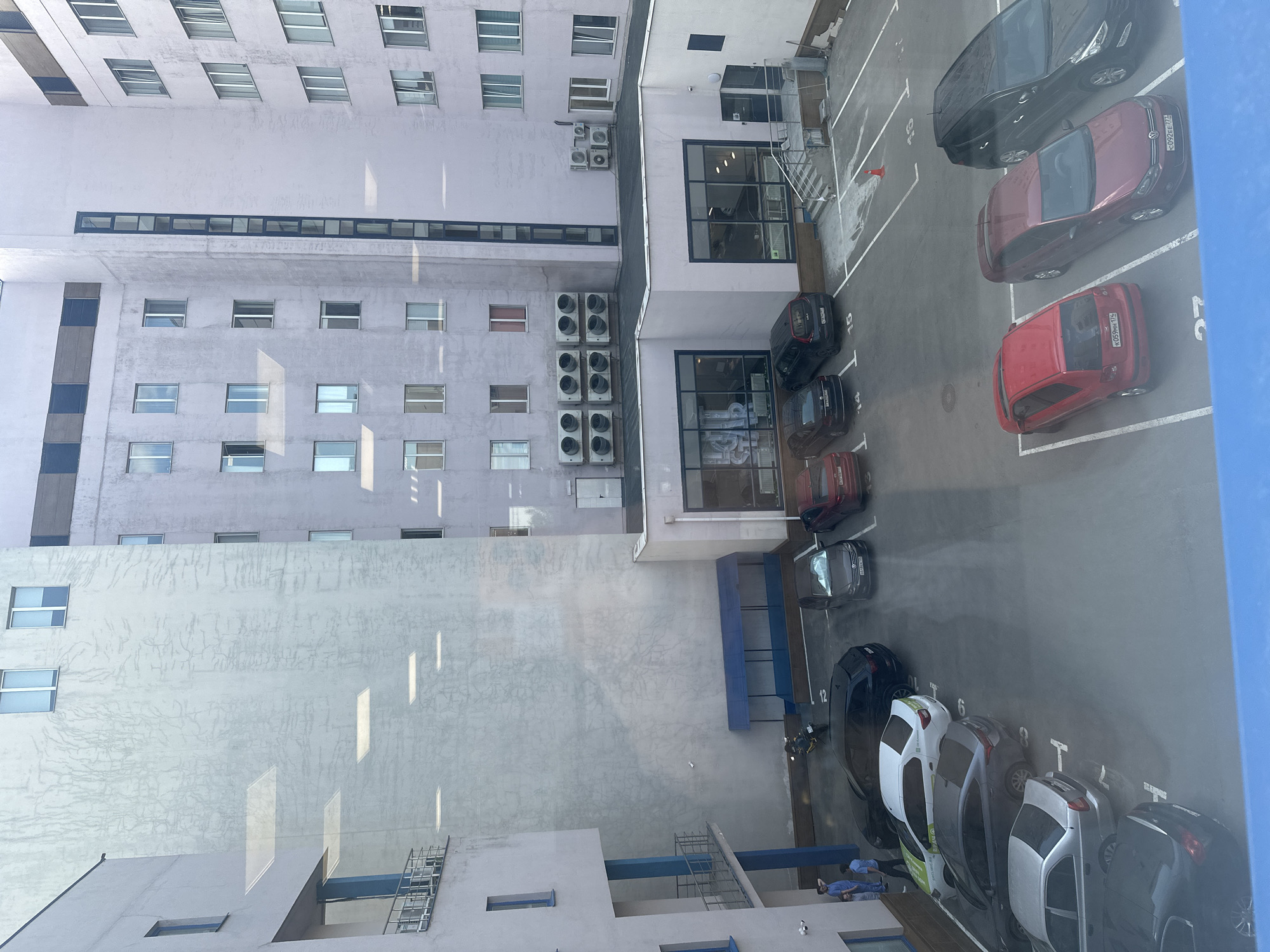 The image depicts an elevated view of a parking lot and adjacent buildings in Moscow, Russia. The buildings are primarily white or light-colored, with a multi-story building featuring rows of windows and external air conditioning units. The parking lot contains several parked cars of various colors, including red, black, white, and gray. Parking spaces are marked with numbers. At least three people are visible near the bottom of the frame, lying down. No discernible actions or interactions are captured in the image. The environment shows a mix of architectural structures and vehicular presence.