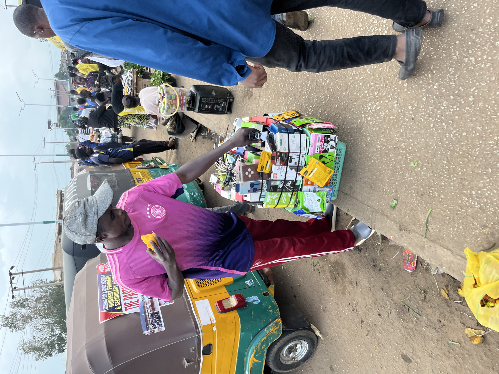 The image captures a busy street scene, likely in Bukuru, Nigeria, as indicated by the context provided. In the foreground, a man wearing a pink and purple soccer jersey and maroon pants stands next to a display of electronic accessories, such as phone covers and headphones, neatly arranged in a green basket. He is holding a piece of fruit and appears to be selecting an item from the display. Beside him is a colorful, three-wheeled vehicle, commonly known as a "keke" or auto-rickshaw. In the background, a bustling market environment is visible, with numerous people, stalls, and other vehicles. The overall atmosphere suggests a vibrant, commercial hub.