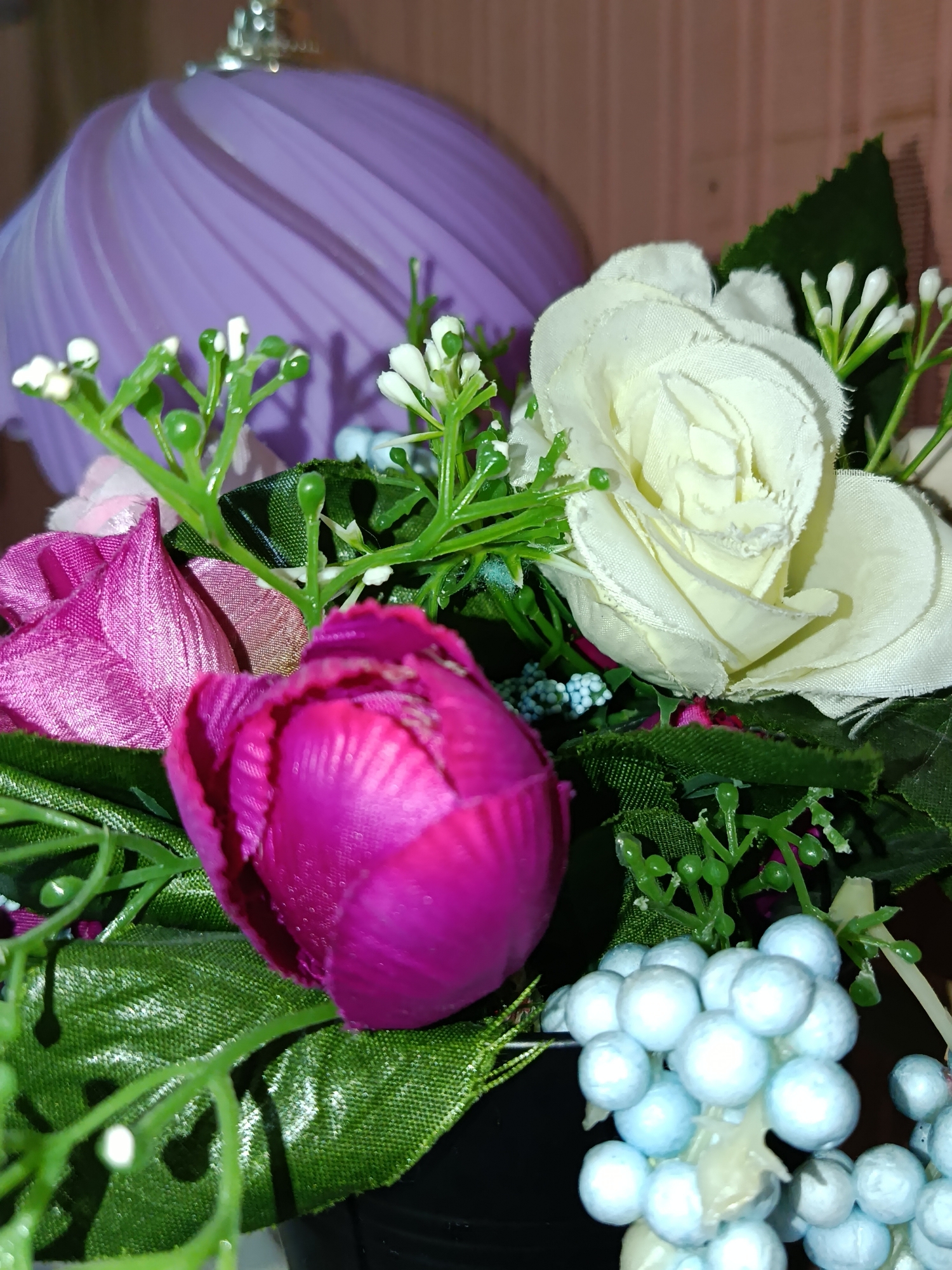 A close-up, eye-level, indoor shot features a bouquet of artificial flowers. The arrangement includes a bright magenta tulip in the foreground, a white rose to its right, and a pink flower to the left. Several small white buds and greenery are interspersed throughout the bouquet. A cluster of light blue, berry-like ornaments is visible at the bottom right. In the blurred background, a portion of a purple, ribbed lamp shade can be seen. The scene is not identifiable as being located in Tashkent, Uzbekistan.