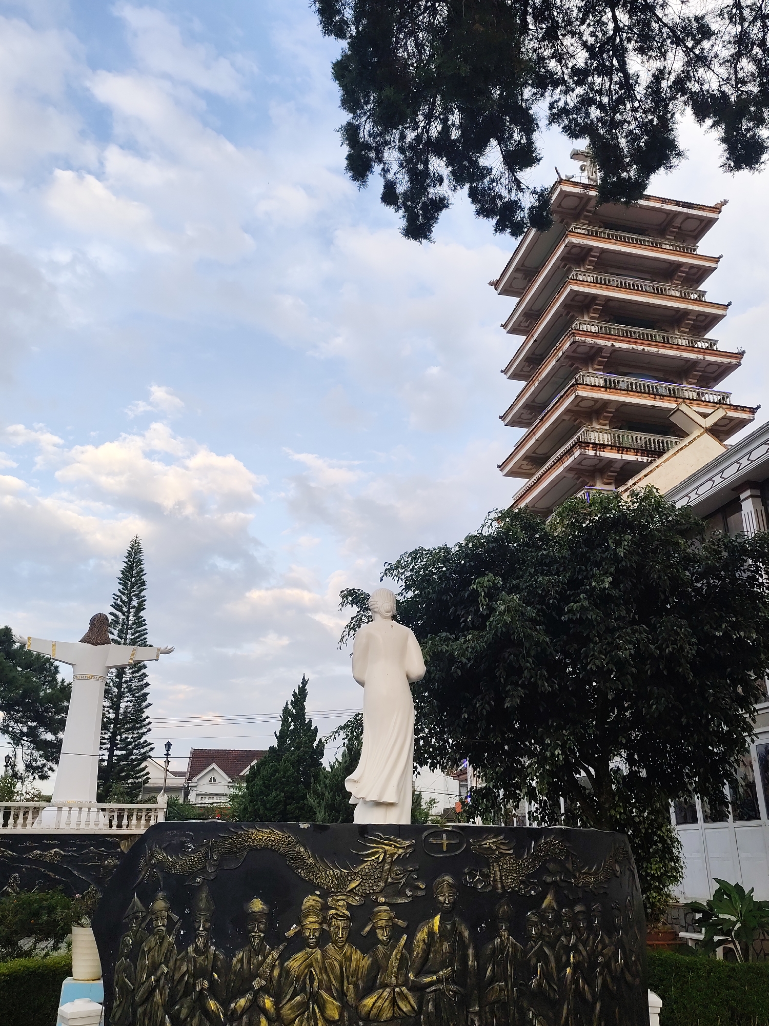 The image shows a view of a multi-story pagoda-like building, a statue of Jesus Christ with open arms, and another white statue of a person facing away from the camera, all situated in a garden or park. The pagoda has a distinct architectural style with multiple tiers and ornate balconies. The statue of Jesus is on a pedestal, and behind it, a tall evergreen tree is visible. In the foreground, a dark relief sculpture depicts a group of figures and a dragon. The scene is set against a sky with scattered clouds, and trees and other buildings are visible in the background. The location is identified as Lâm Viên - Đà Lạt, Vietnam.