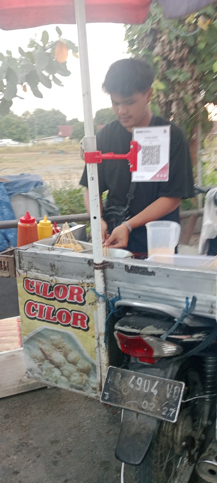 A young male stands behind a food cart selling "Cilor," a type of Indonesian street food, in Ngemplak, Indonesia. The cart is mounted on a motorcycle, indicated by the visible license plate "AB 4904 VO" and rear fender. The cart itself is made of metal and features a prominent "CILOR CILOR" sign. Two bottles of condiments, likely chili sauce and mustard, are visible on the cart. The young man is wearing a black t-shirt and appears to be in the process of preparing the food, holding skewers of what seem to be fried dough balls. A QR code for payment is attached to a red bracket on a white pole supporting the cart's awning. The background shows a blurry outdoor environment with trees and some distant buildings. The lighting suggests it is either early morning or late afternoon.