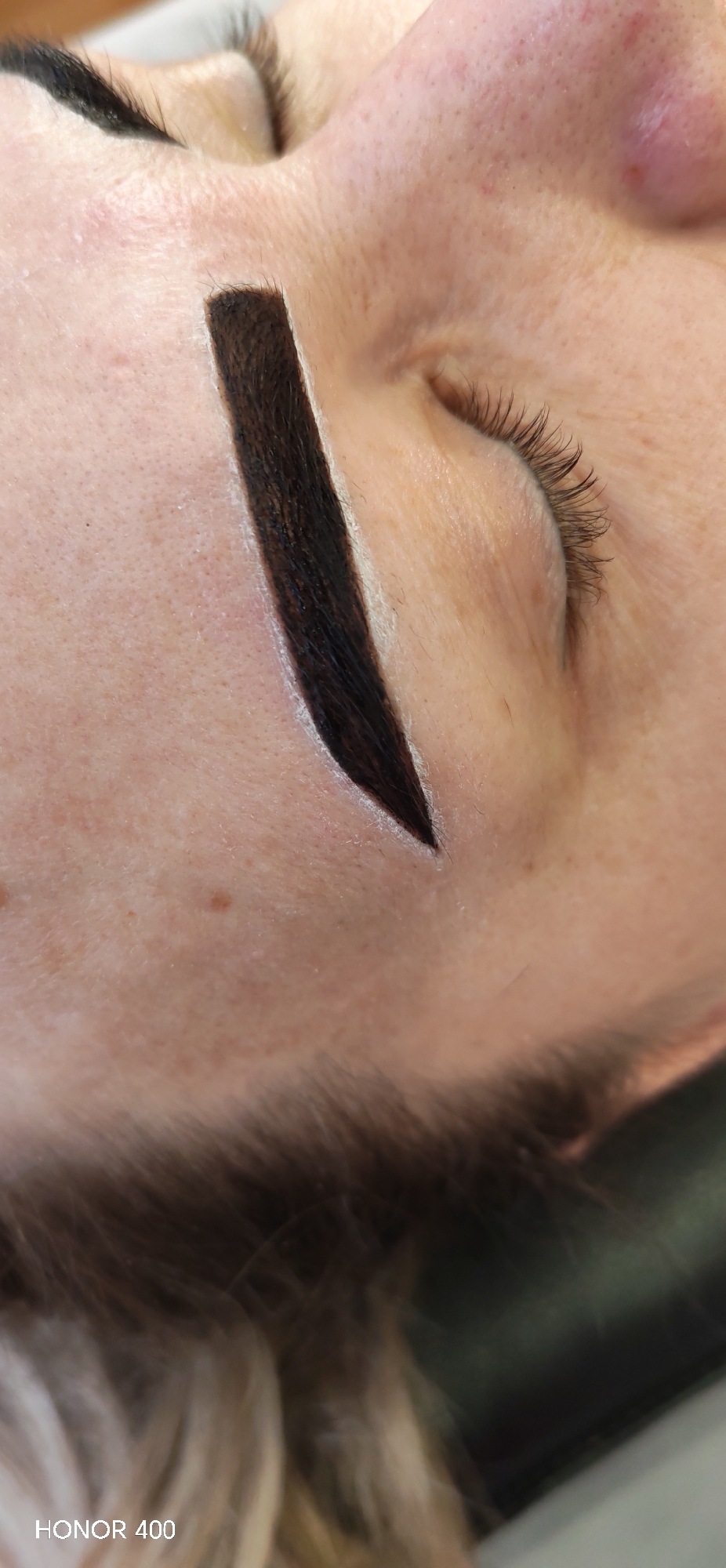 A close-up, high-angle vertical shot captures the left side of a human face, likely female, with the eye closed. The subject's left eyebrow is undergoing a cosmetic procedure, with a dark brown or black pigment applied, precisely outlined by a white marking paste. The eyebrow shape is defined with a broad front and a sharply tapered tail. Long, dark eyelashes are visible on the closed eyelid. The fair skin exhibits visible pores and minor blemishes. Light-colored hair is blurred at the bottom of the frame. The background is indistinct and dark. An 'HONOR 400' watermark is present in the lower left. The specific location within the city of Chaikovsky, Russia, is not discernible from the image.