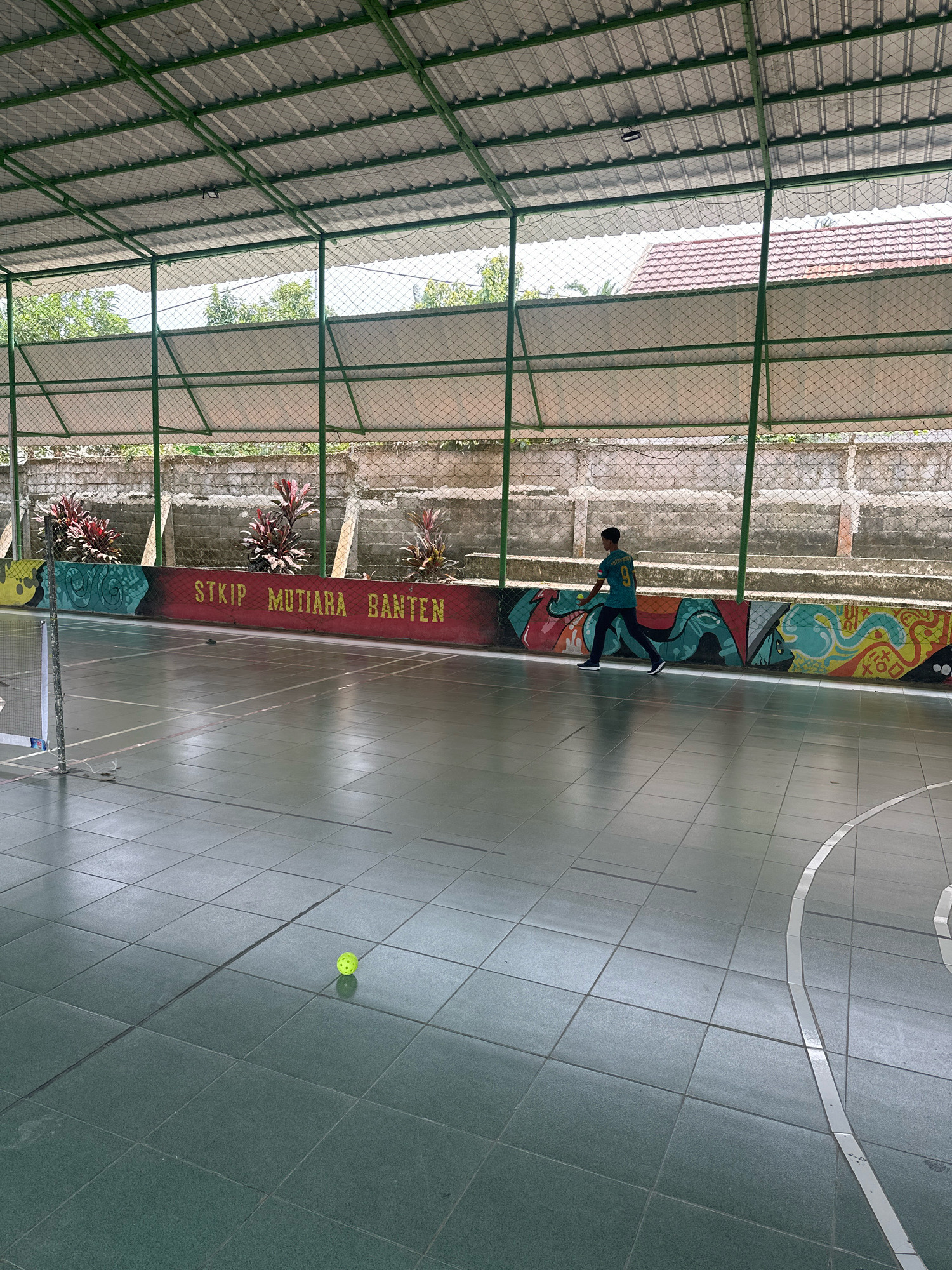 A young boy wearing a blue t-shirt with the number 91 on the back is walking across a green tiled indoor court. He is holding a yellow ball in his left hand, near his waist. The court is enclosed by a green metal fence and covered by a corrugated metal roof supported by green metal beams. Along the back wall of the court, there is a red mural with yellow text that reads "STKIP MUTIARA BANTEN," and colorful graffiti art. The background shows a brick wall and some greenery. The image appears to be taken in Saruni, Indonesia.