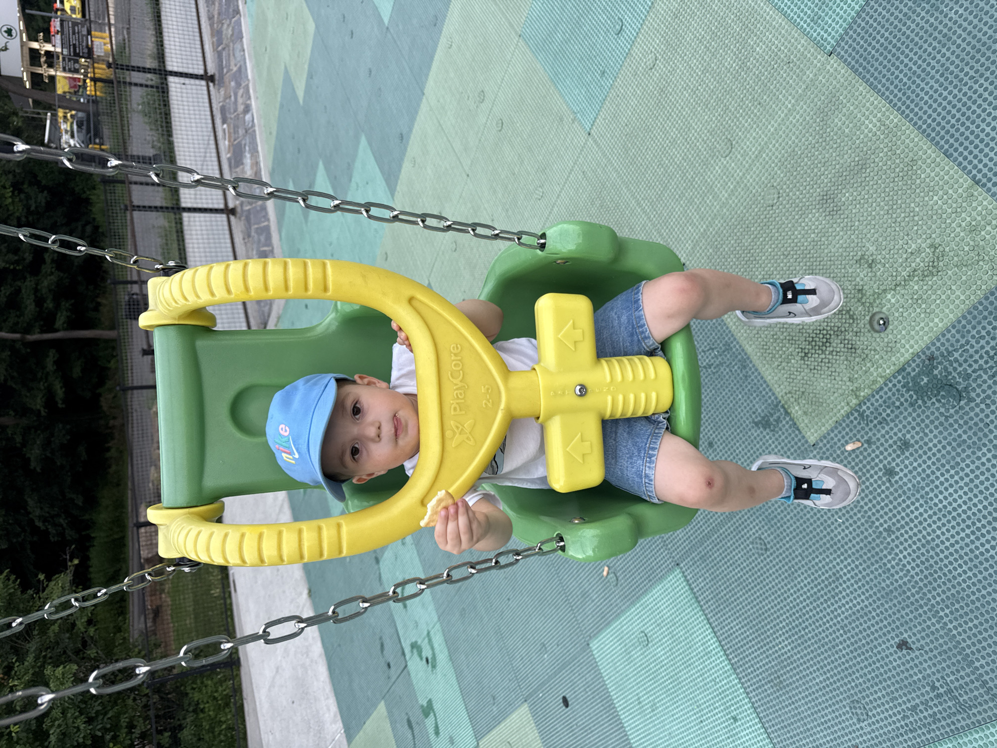 A young child, wearing a blue baseball cap and denim shorts, sits in a green and yellow plastic swing. The child is holding a piece of food in their right hand and their tongue is sticking out. The swing is suspended by metal chains and is located in a playground with a patterned rubber mat surface. In the background, trees and a fence are visible, with buildings and vehicles in the distance, suggesting an urban environment. The image is taken outdoors during daylight.