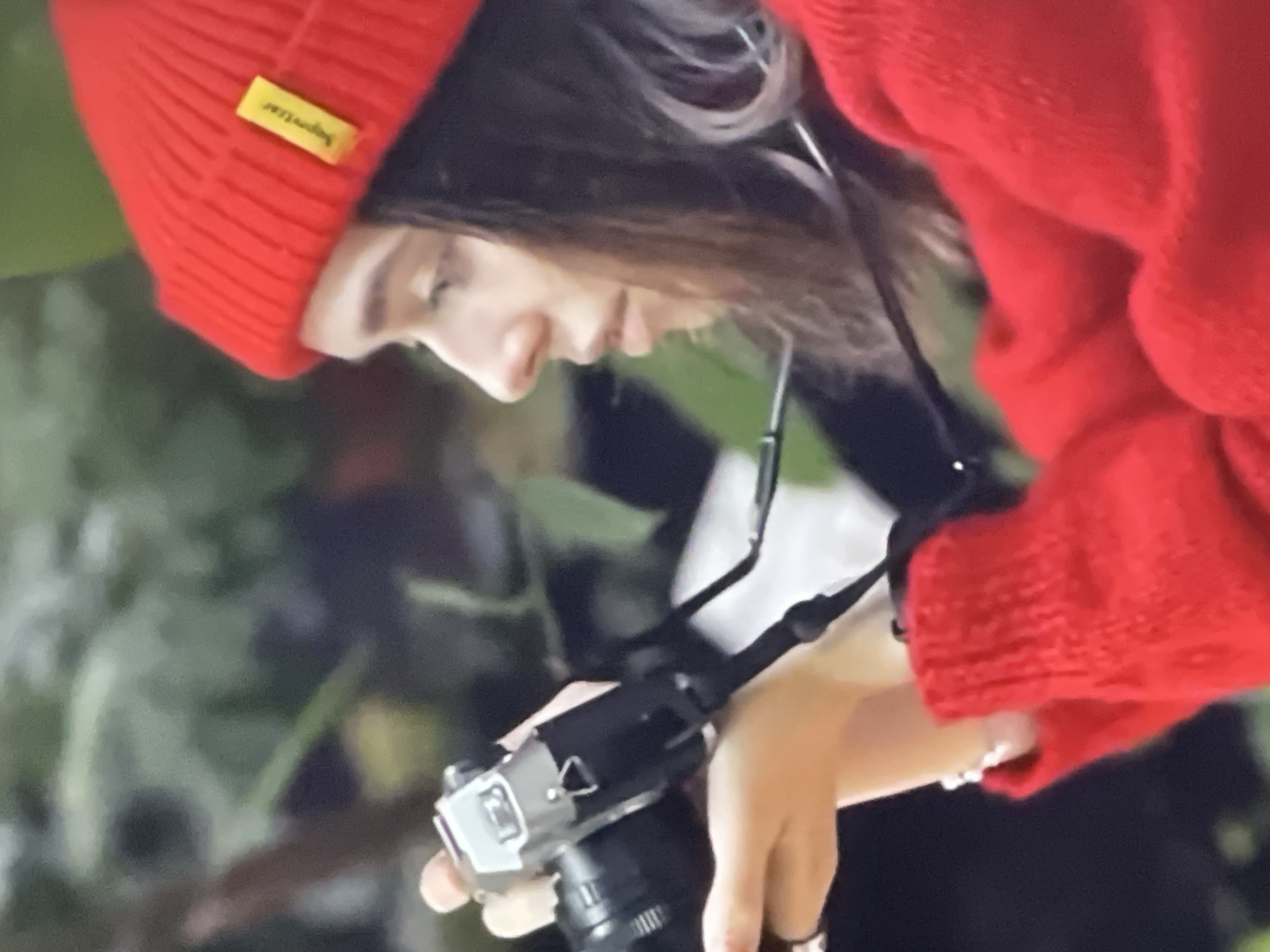 A woman wearing a red beanie and red sweater is holding a silver camera and appears to be taking a picture in an outdoor setting. The background consists of blurred green foliage. The image does not contain any information that would suggest the location is Pattaya City, Thailand.