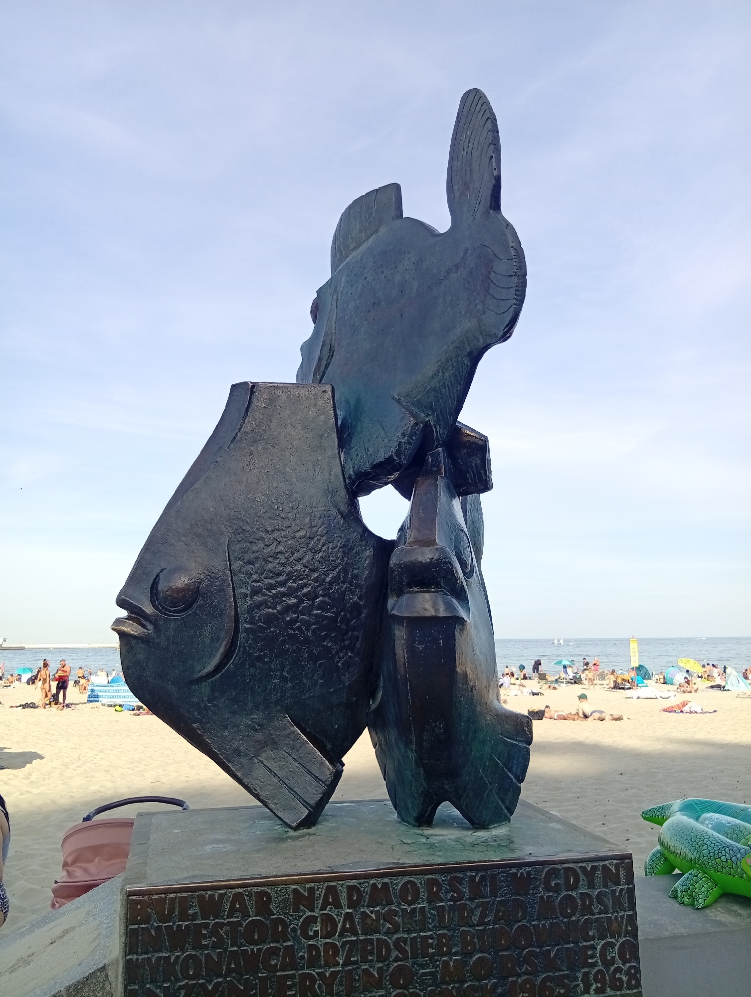 The image shows a large, dark metal sculpture of two fish-like creatures on a sandy beach in Gdynia, Poland. The sculpture is positioned on a concrete base with a bronze plaque at its front. The plaque contains text, partially obscured by the sculpture, but it appears to list names and dates. In the background, the Baltic Sea is visible with a few sailboats on the water. The beach is populated with numerous sunbathers, beach umbrellas, and tents. To the right of the sculpture, a large, inflatable green turtle is partially visible. The sky is clear and blue. The sculpture's design is abstract and geometric, with sharp edges and stylized features. The texture of the metal is rough and appears weathered. The overall scene depicts a public art installation in a recreational area.